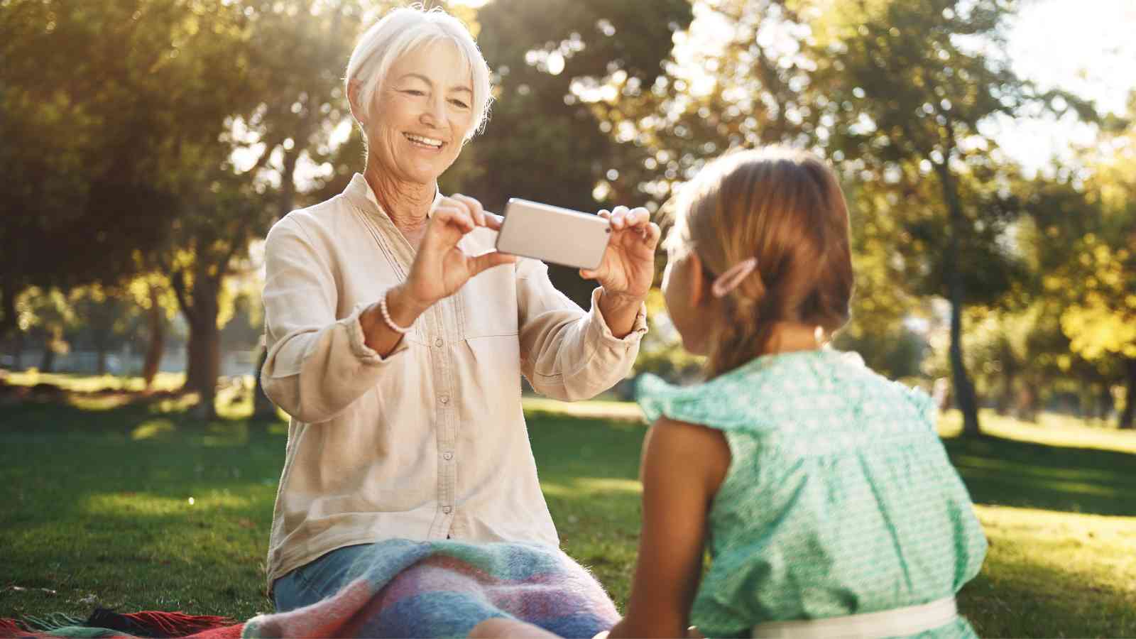 An older woman takes a photo of a young girl sitting in front of her on a blanket in a sunlit park.
