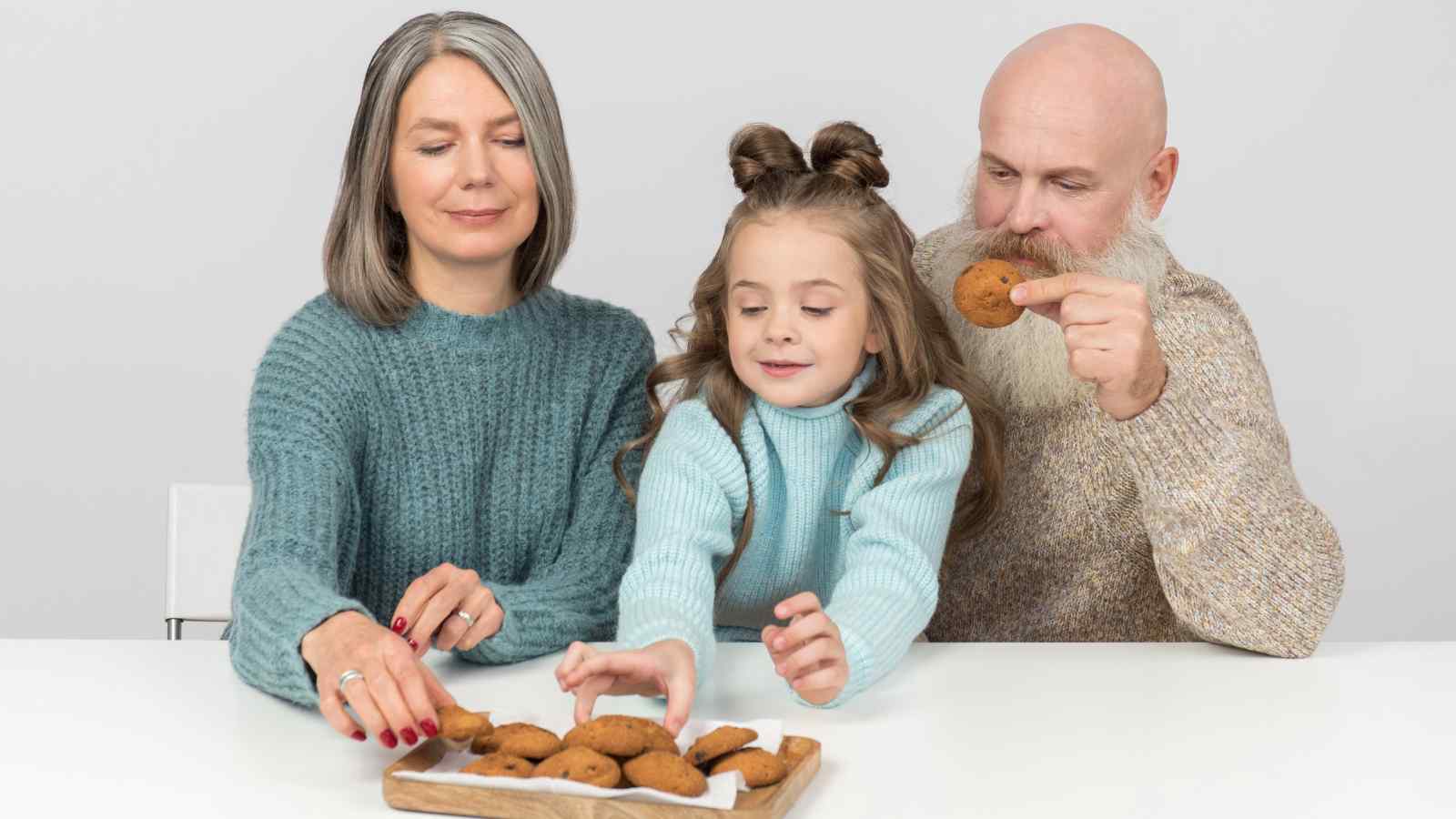Three people, two adults and a child, sit at a white table reaching for and eating cookies from a tray.