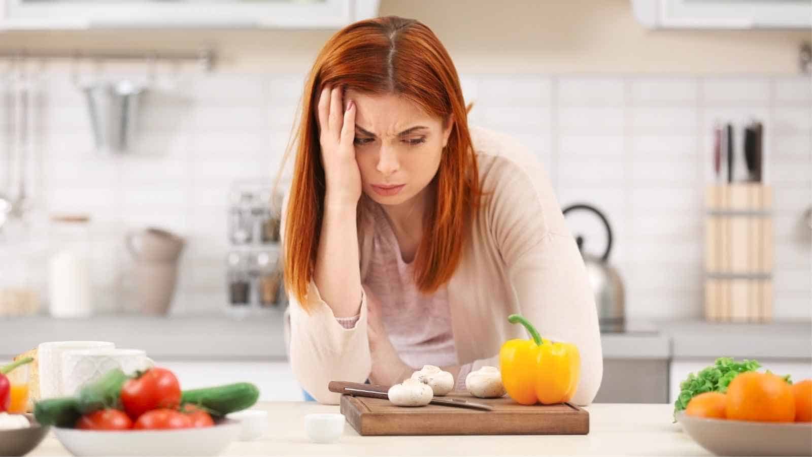A woman with red hair looks stressed while preparing vegetables, including mushrooms and a yellow bell pepper, on a cutting board in a kitchen.