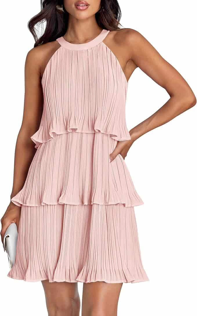Woman wearing a sleeveless, light pink, pleated tiered dress, holding a small white clutch in her left hand.