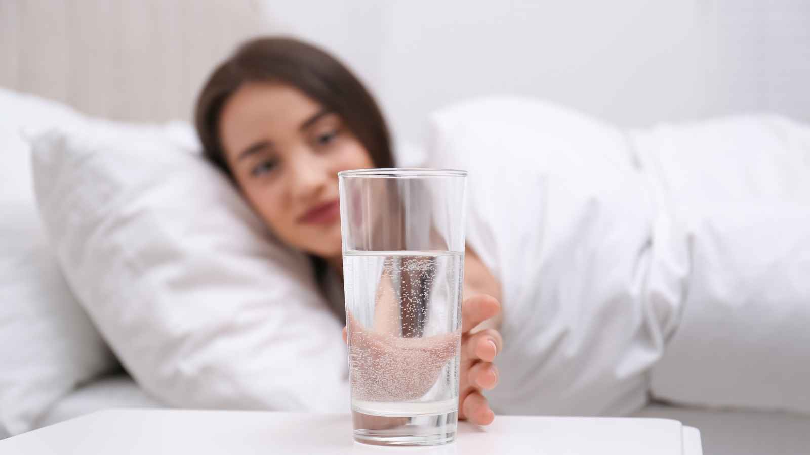 A woman lying in bed reaches for a glass of water on a bedside table, with white bedding in the background.
