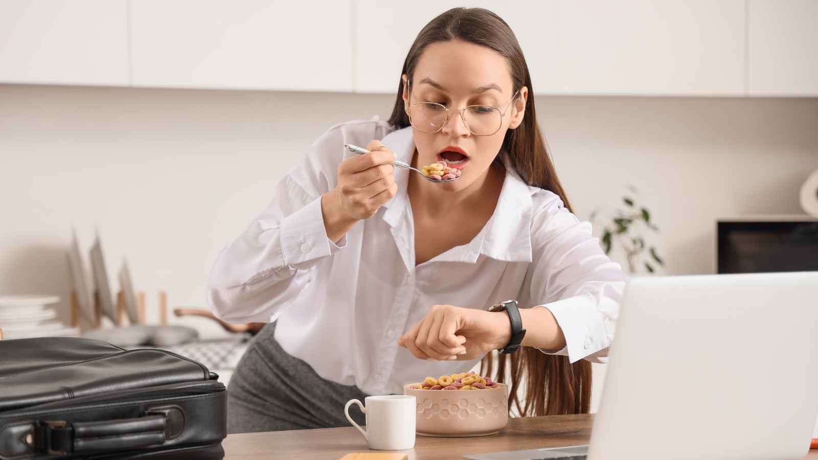 A woman in business attire quickly eats cereal while looking at her wristwatch, with a laptop and briefcase on the table in a kitchen setting.