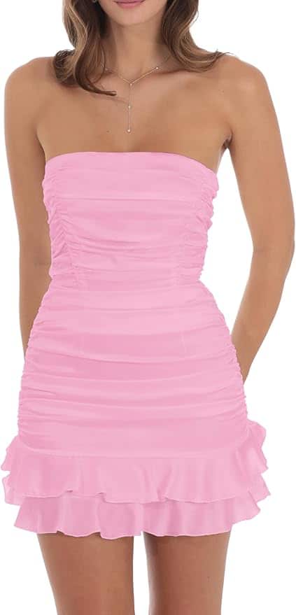 A woman wearing a strapless, ruched light pink mini dress with a ruffled hem, standing with her hands behind her back.