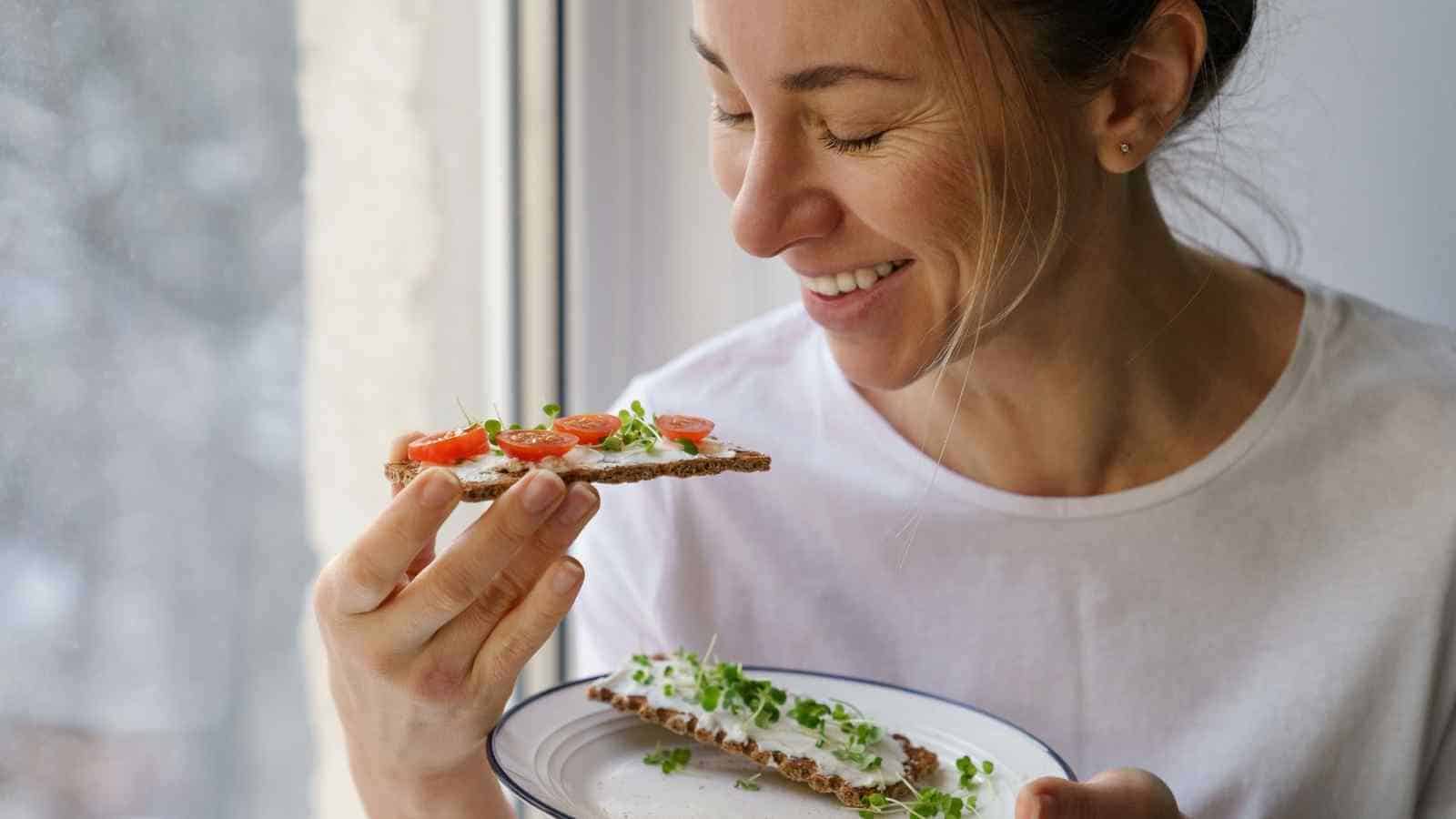 A woman holding a plate and smiling at a crispbread topped with cream cheese, tomato slices, and greens near a window.
