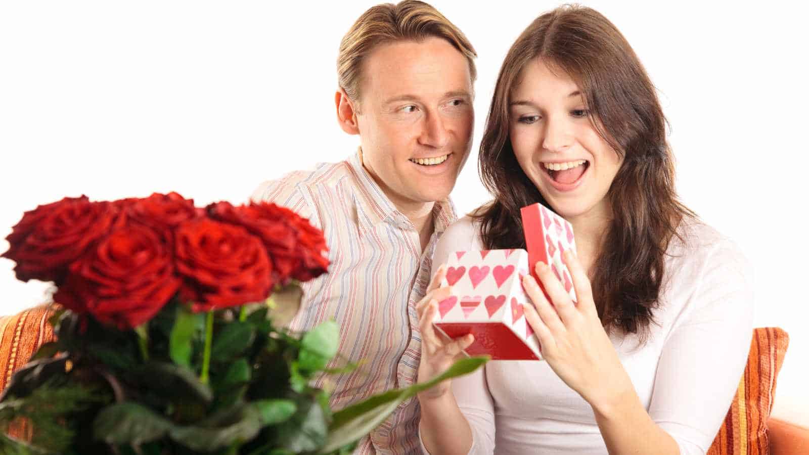 A woman opens a heart-patterned gift box and smiles while a man sits beside her. A bouquet of red roses is in the foreground.