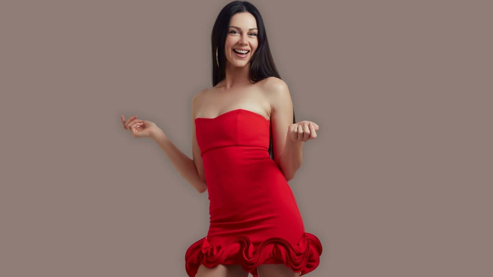 A woman in a strapless red dress with a ruffled hem stands smiling against a plain taupe background.