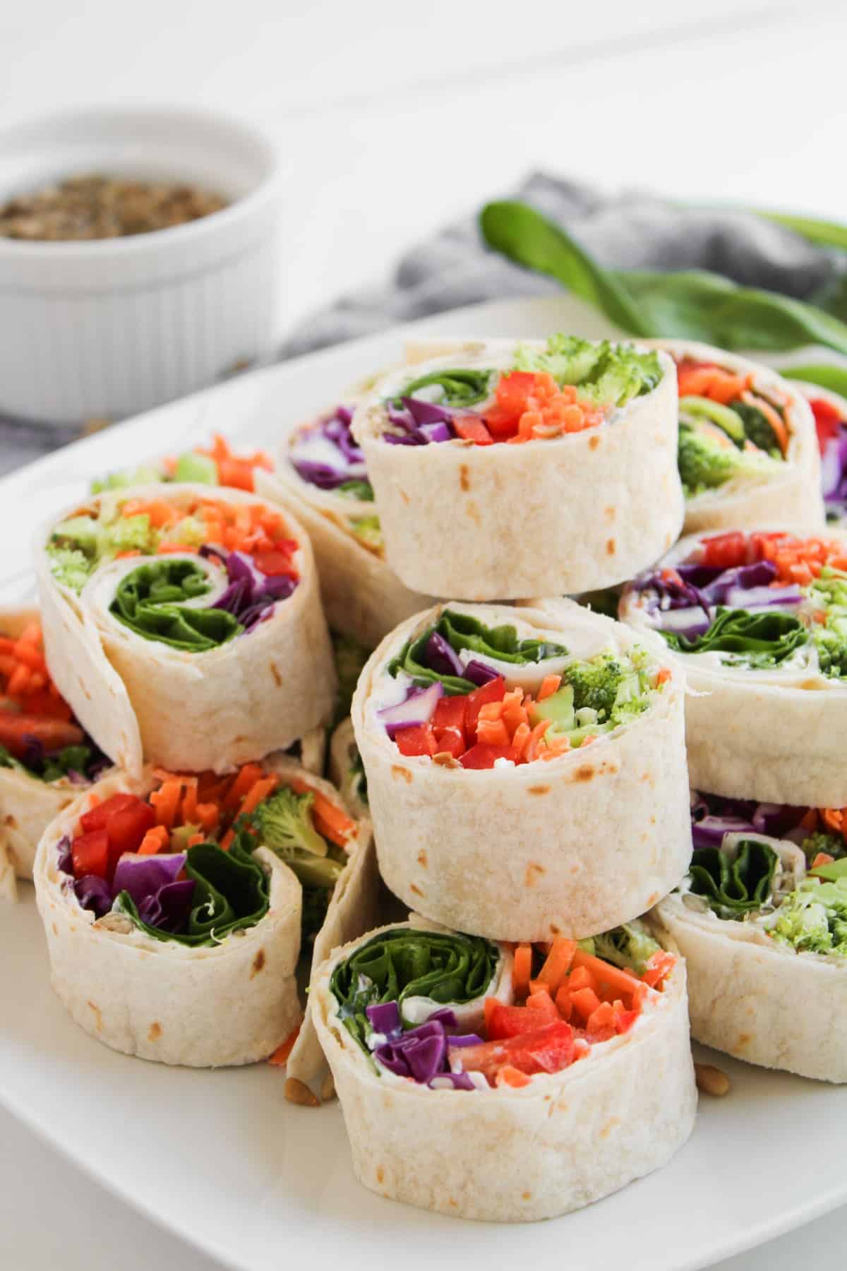 A plate of tortilla pinwheel wraps filled with colorful vegetables such as spinach, carrot, red bell pepper, and purple cabbage, stacked on a white surface.