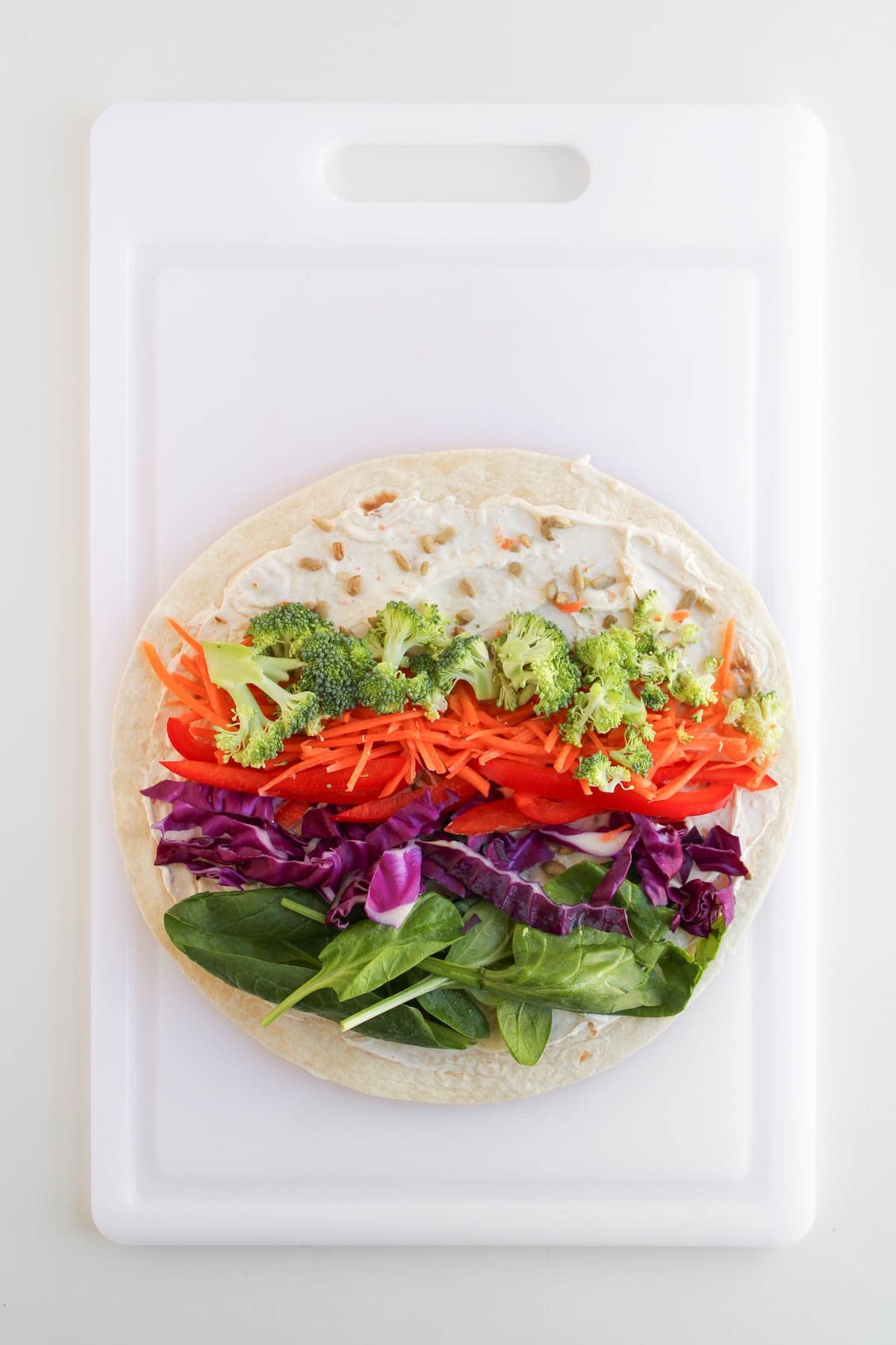 A tortilla wrap on a cutting board topped with spinach, purple cabbage, shredded carrots, red bell pepper, broccoli, and a spread.