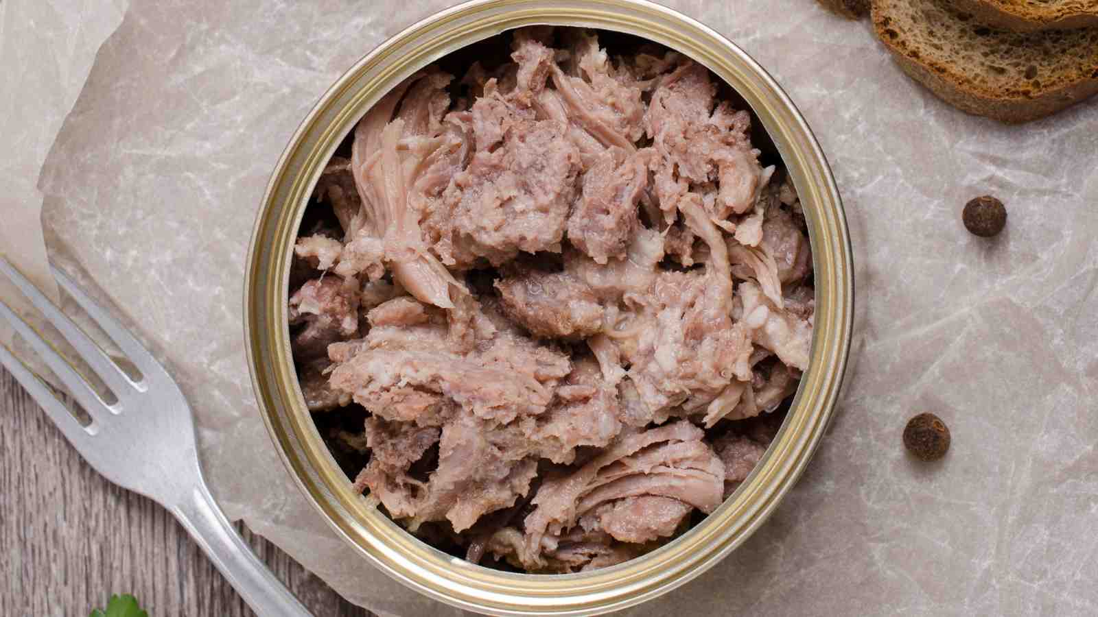 An open can of shredded tuna or meat, placed on parchment paper with a fork, slices of bread, and peppercorns nearby.