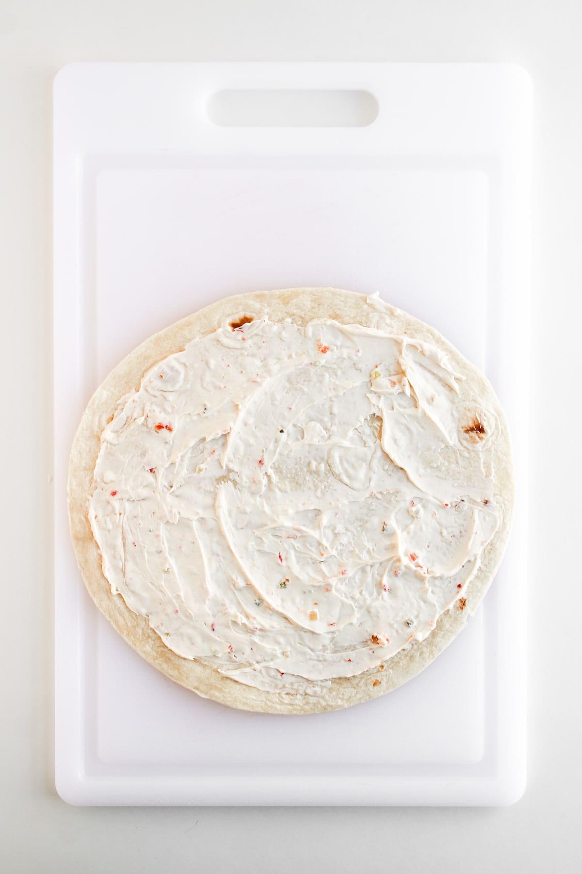 A flour tortilla with a layer of creamy white spread on top, placed on a white cutting board.