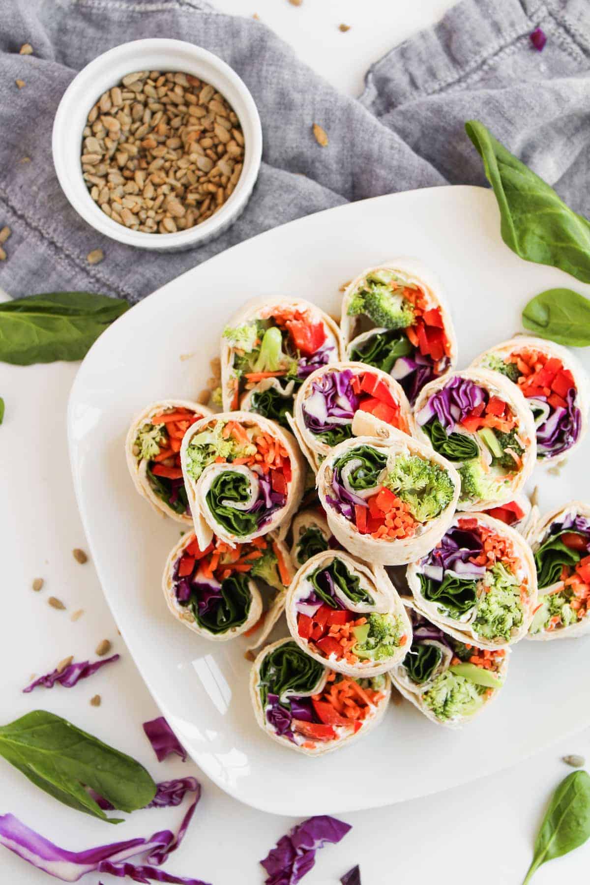 A plate of vegetable wraps filled with spinach, carrots, red cabbage, and broccoli, surrounded by loose greens, with a small bowl of seeds in the background.