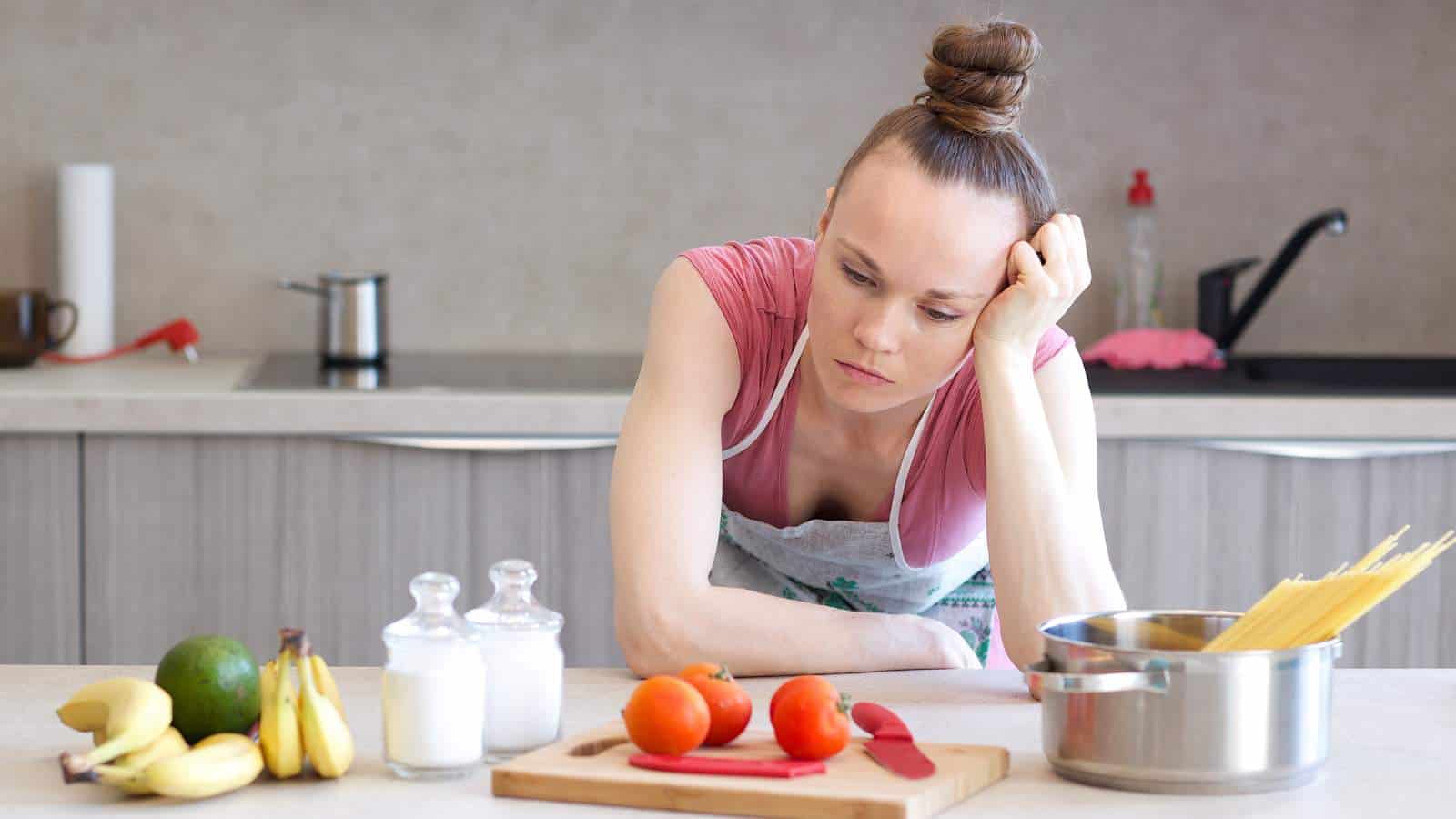 A woman in a kitchen leans on the counter, looking at tomatoes on a cutting board, with uncooked spaghetti, fruit, salt, and sugar nearby.