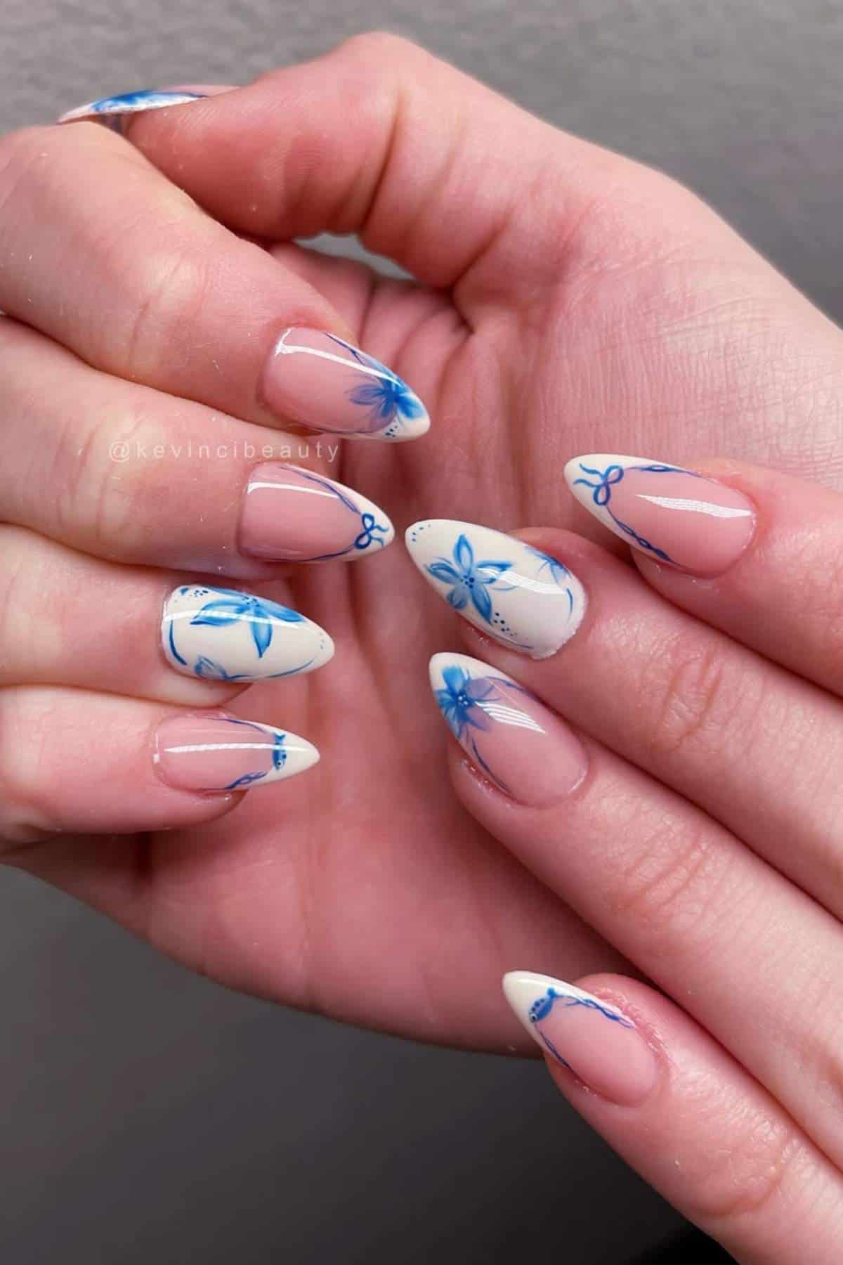 A close-up of hands with almond-shaped nails featuring a nude base, white tips, and blue floral designs on each nail.