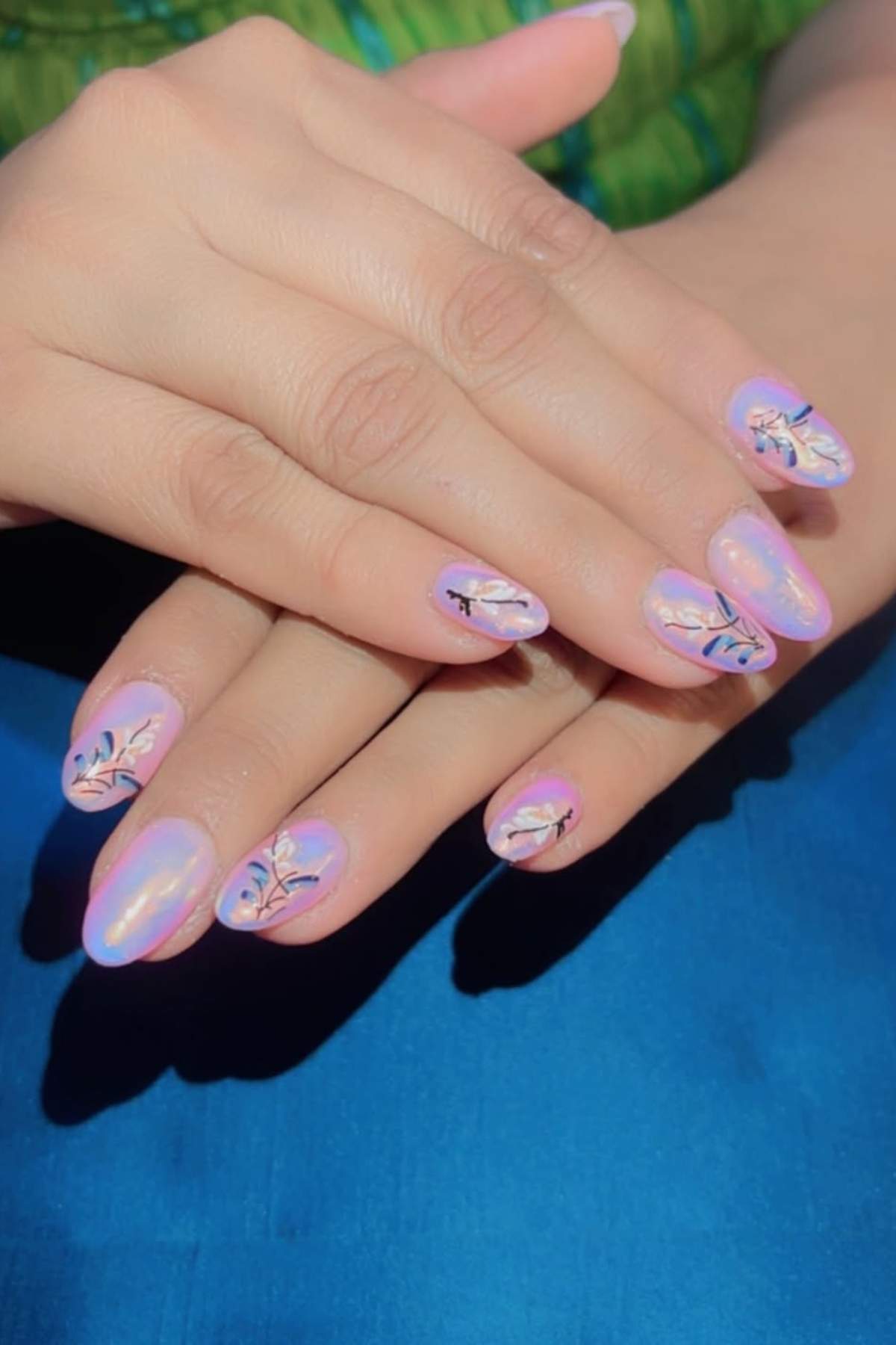 Hands with long, almond-shaped nails painted light pink and decorated with delicate black and white floral designs, resting on a blue and green background.