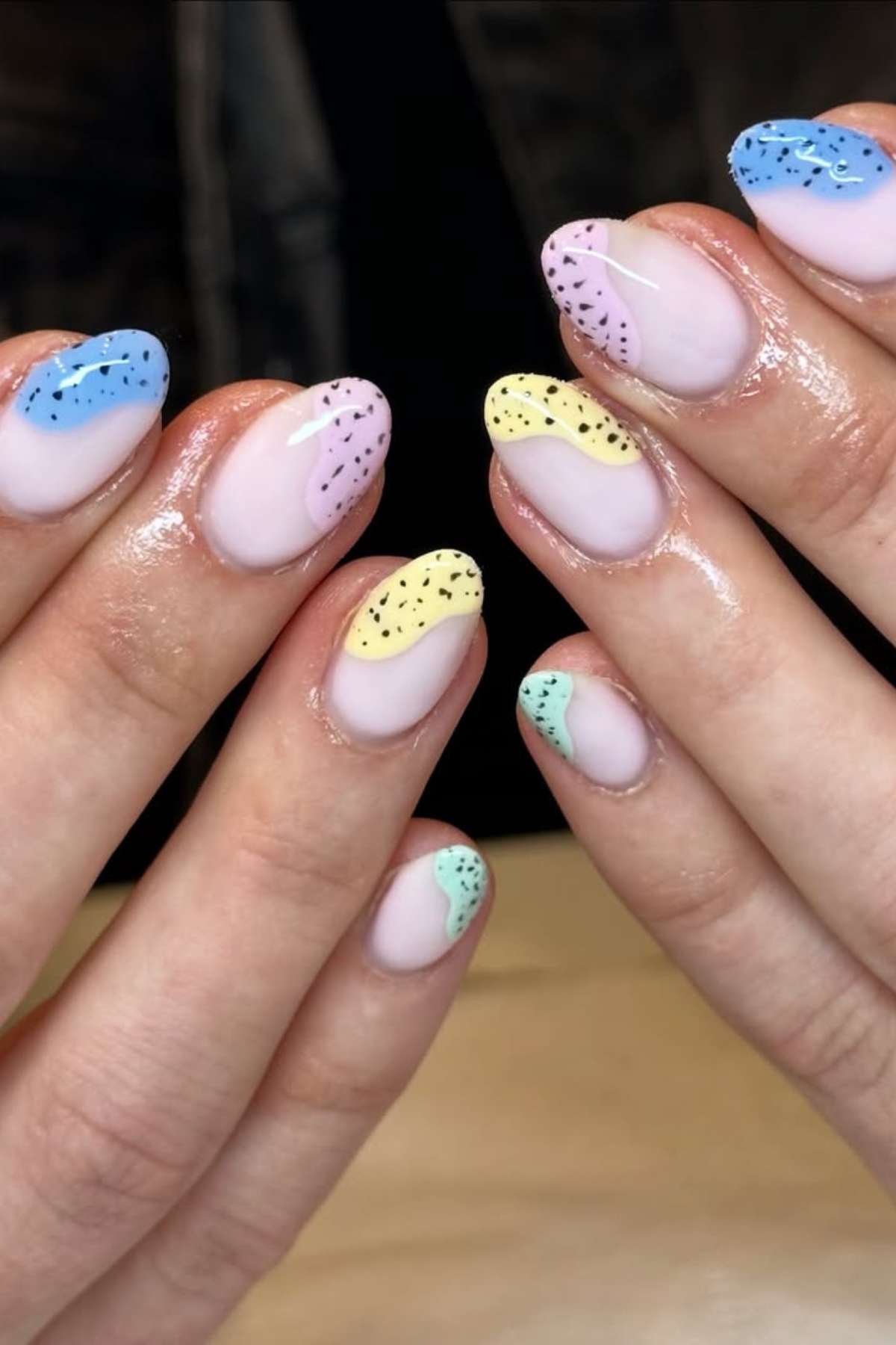 Hands with almond-shaped nails featuring pastel French tips in blue, yellow, pink, and green, each accented with small black speckles.