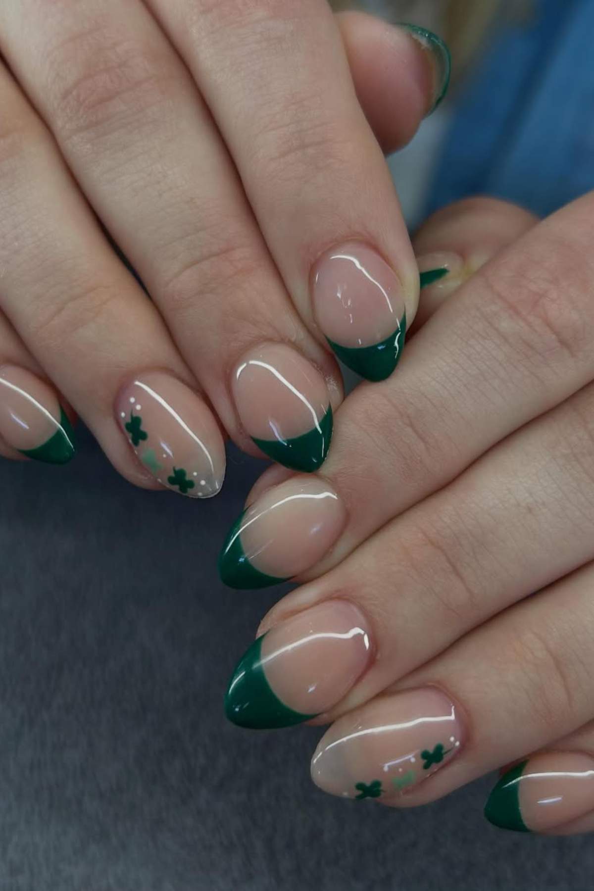 Close-up of hands with almond-shaped nails featuring green French tips and small green shamrock designs on a nude base.