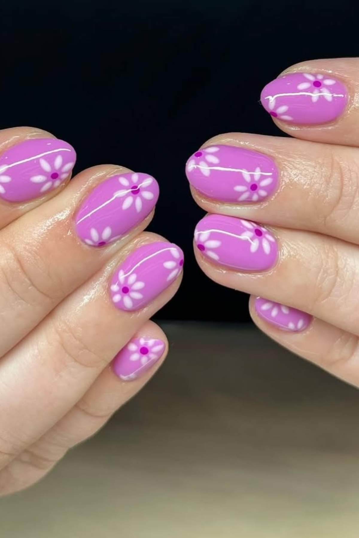 Hands with short, rounded nails painted purple, decorated with white daisy flower designs and small purple dot centers on each nail.