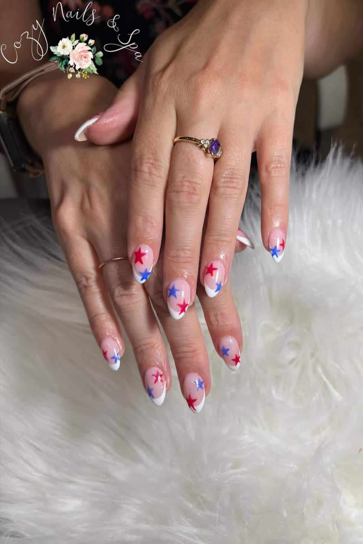 Hands with almond-shaped nails featuring a white base and red and blue star designs, resting on a white fur surface. A gold ring with a purple stone is worn on one finger.