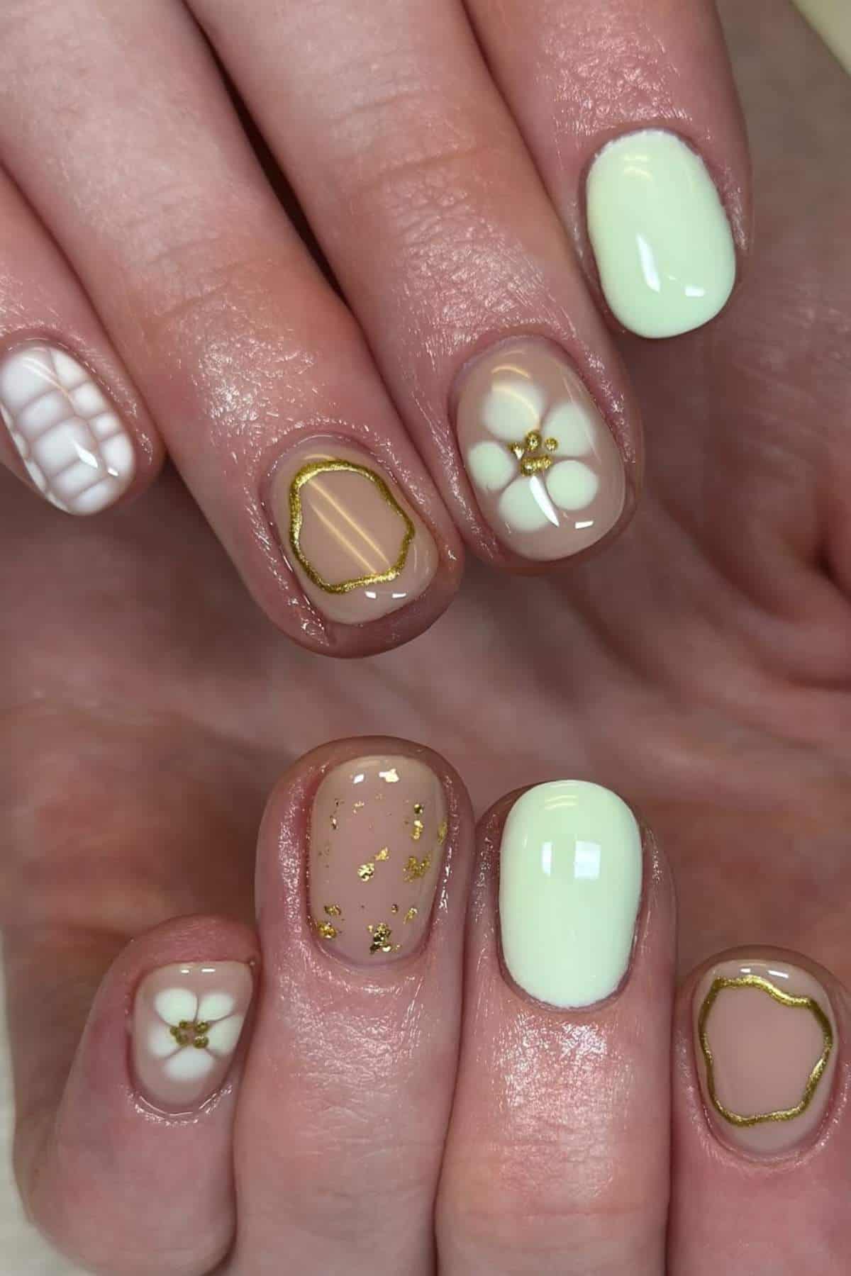 Close-up of hands with manicure featuring pastel green, nude, and white nails, gold accents, floral designs, and a white grid pattern on one nail.