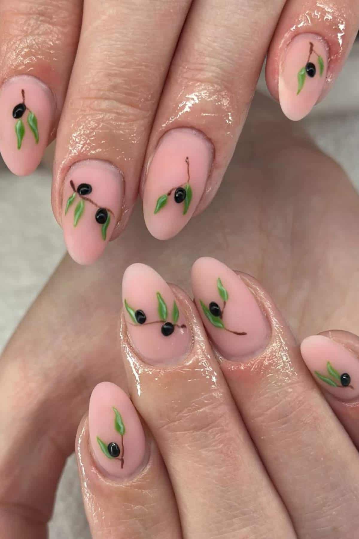 Hands with almond-shaped nails, painted in a nude pink shade with small black dots and green leaf designs resembling olives on each nail.