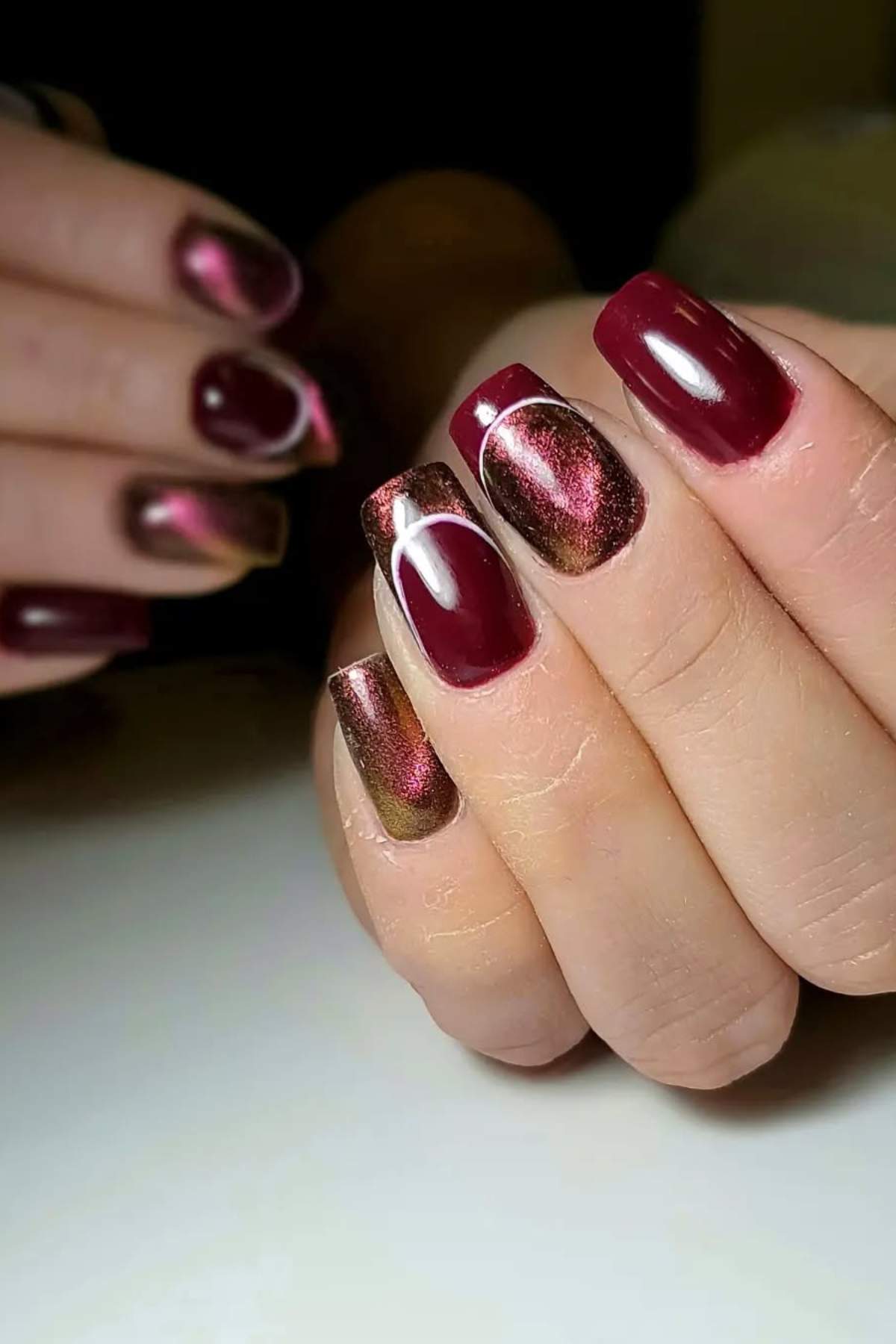 Close-up of a hand with short, square-shaped nails painted in a shiny burgundy and metallic red gradient design, with accent nails featuring a cat-eye effect.