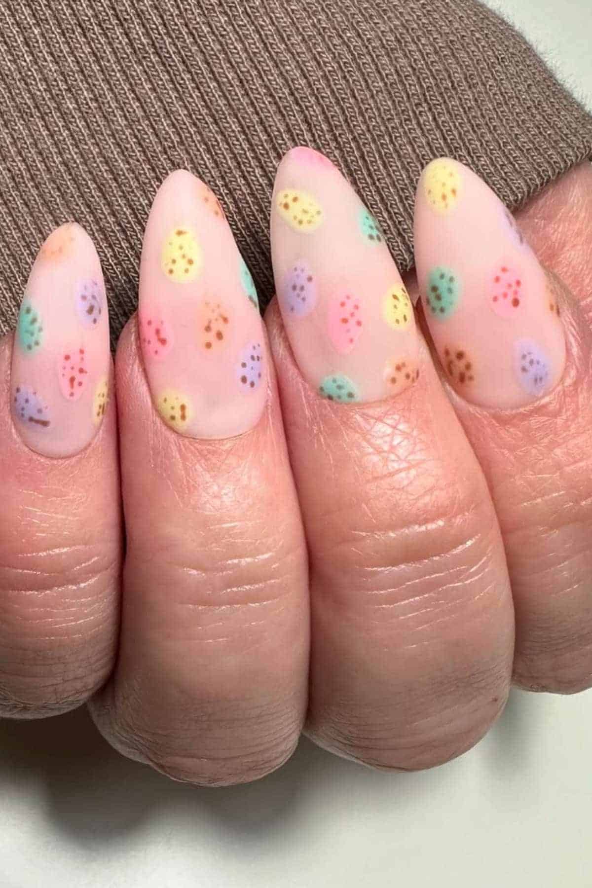 A hand with almond-shaped nails painted in a matte pale pink polish, decorated with small multicolored polka dots resembling Easter eggs.