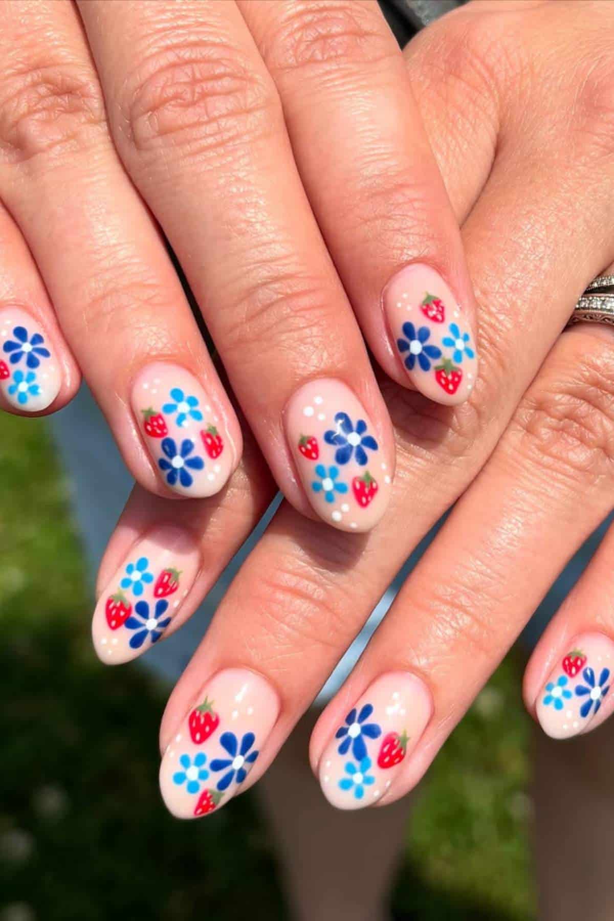 Hands with almond-shaped nails featuring blue flowers and small red strawberries painted on a light nude base, shown outdoors in natural light.