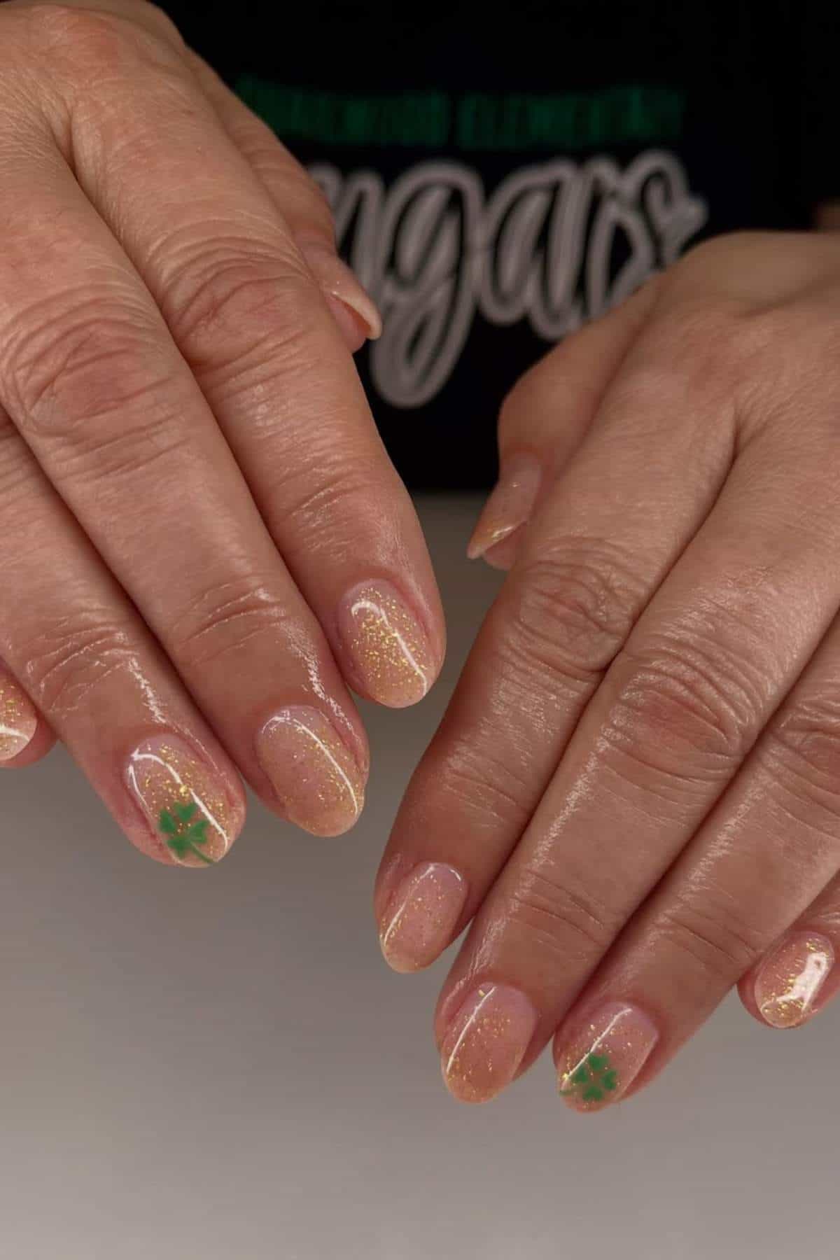 Two hands with short, rounded nails feature a shimmery nude polish and small green four-leaf clover designs on a few nails.