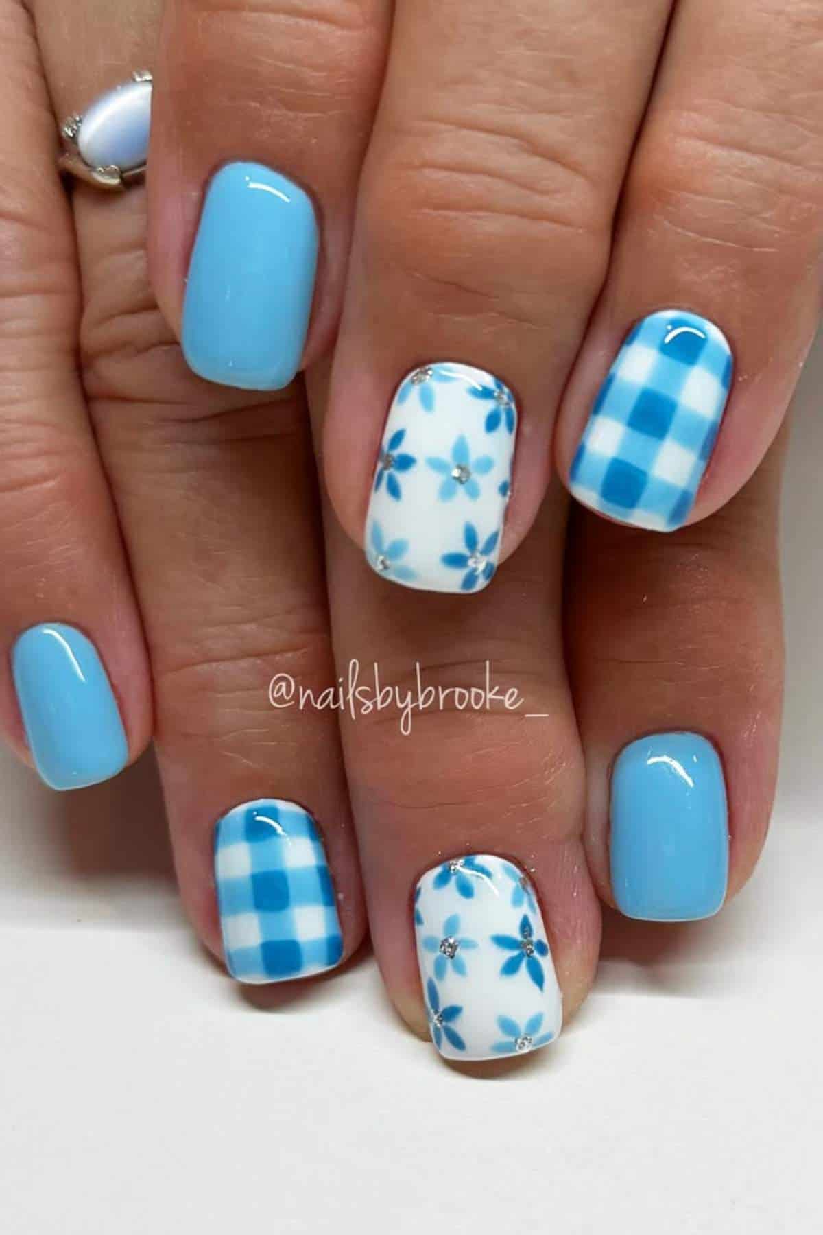 Hands with short, square-shaped nails featuring light blue polish, blue and white checkered patterns, and blue floral designs on two accent nails.