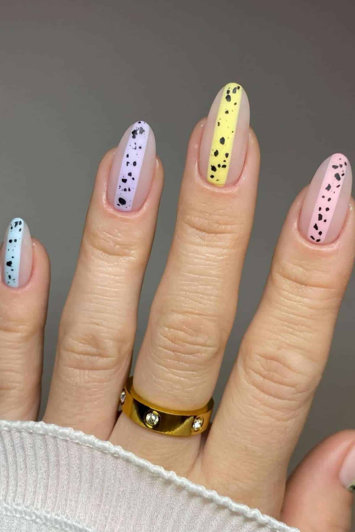 A hand with pastel-colored nails in blue, purple, yellow, and pink, each featuring black speckled dots. The person wears a gold ring with small stones.