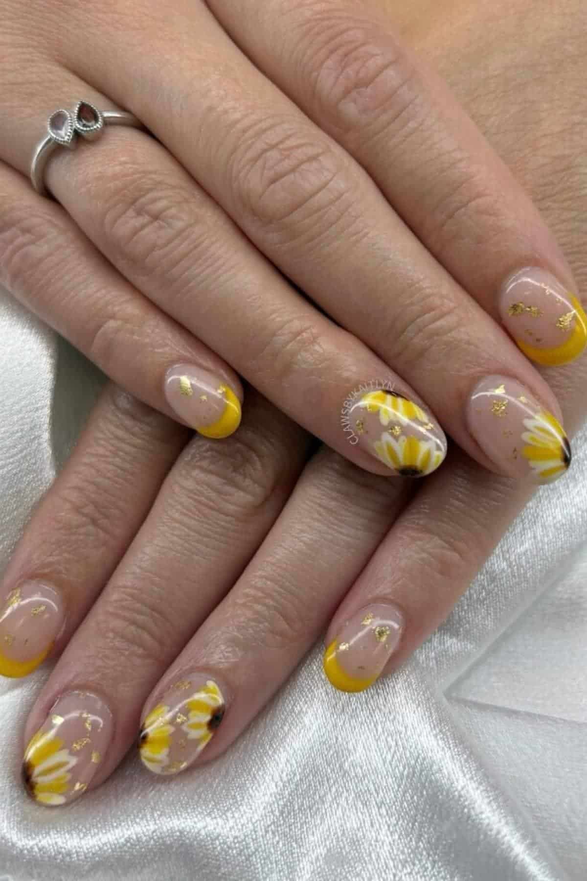 Hands with short, almond-shaped nails featuring yellow French tips and daisy flower nail art accented with small gold flakes. The person is wearing a silver ring.