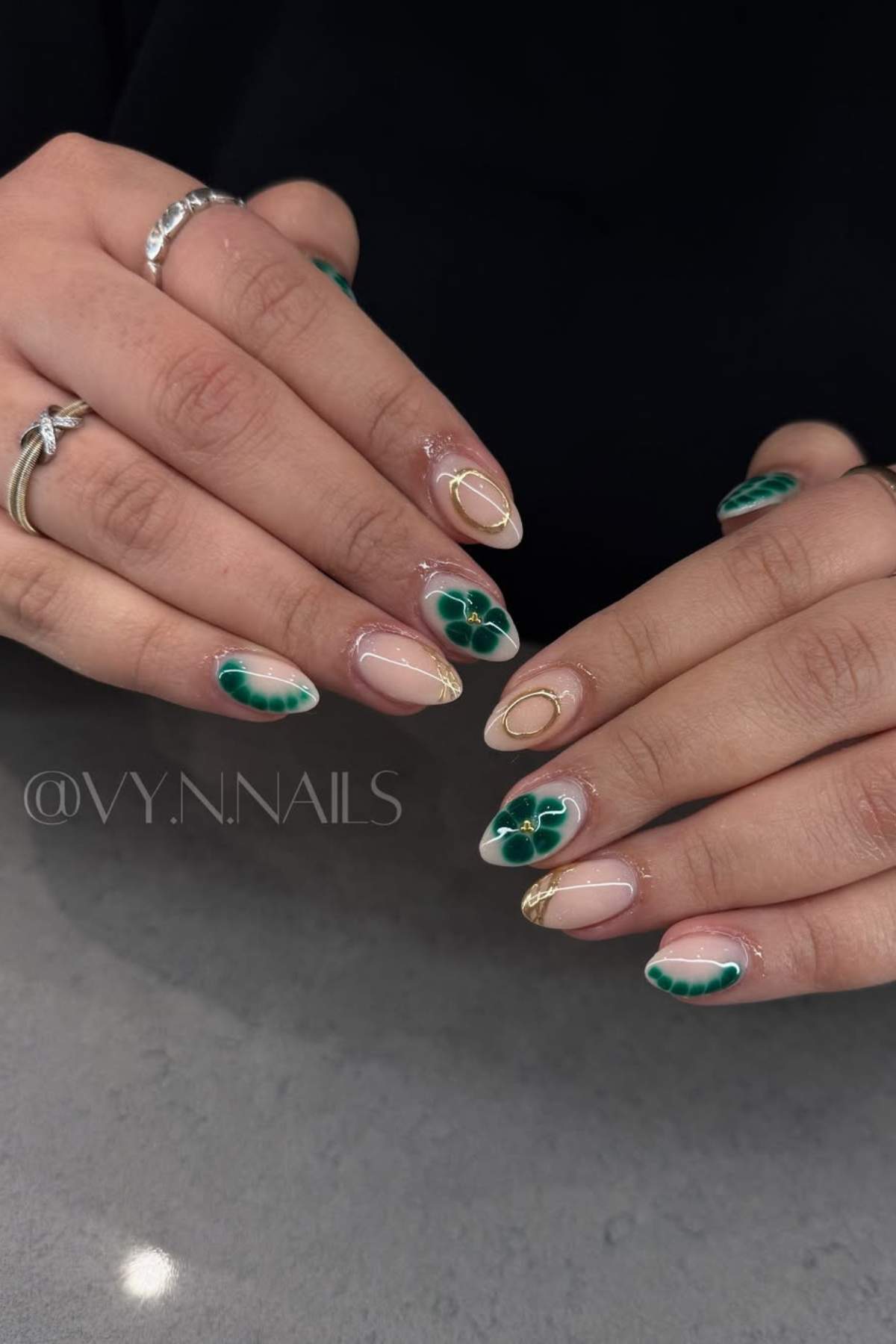 Hands with almond-shaped nails featuring green marble designs, gold accents, and nude bases, displayed on a gray surface.