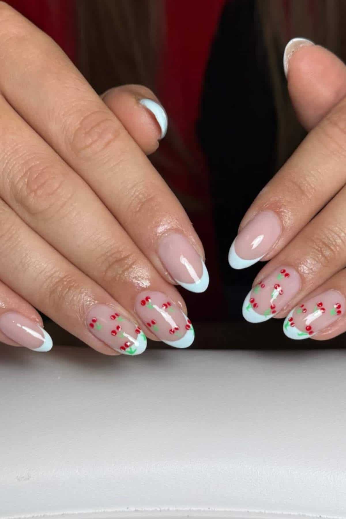 Hands with almond-shaped nails; some nails have white French tips, others have small red and green cherry designs on a nude base.