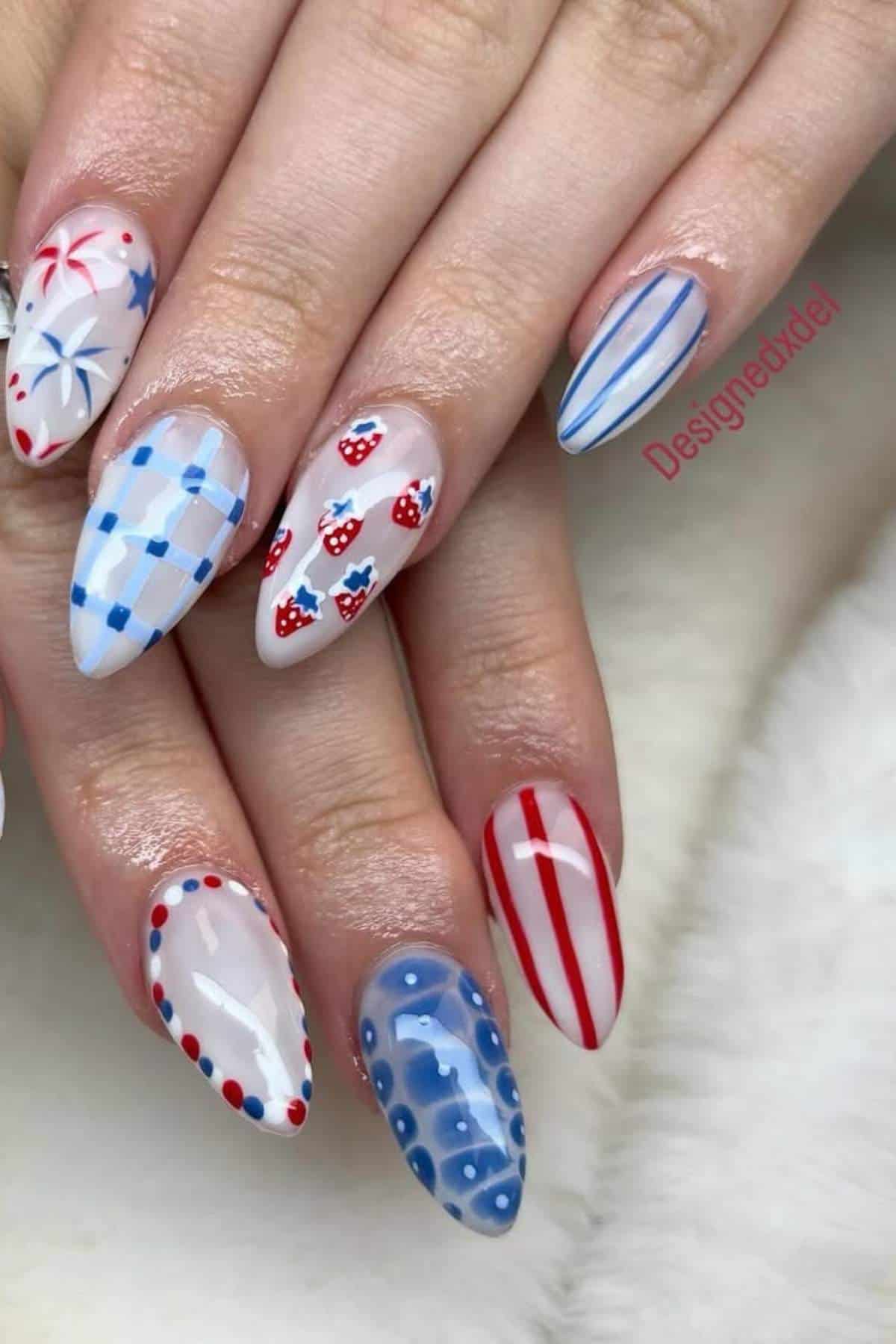 A hand displays almond-shaped nails with various red, white, and blue designs, including stripes, checks, dots, stars, and small cherries on a white background.