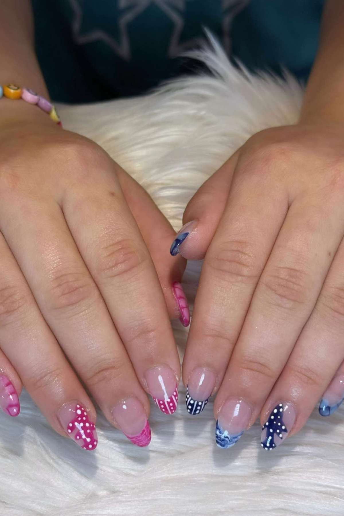 Two hands with manicured nails featuring various pink, blue, and white patterned designs on a white furry surface.