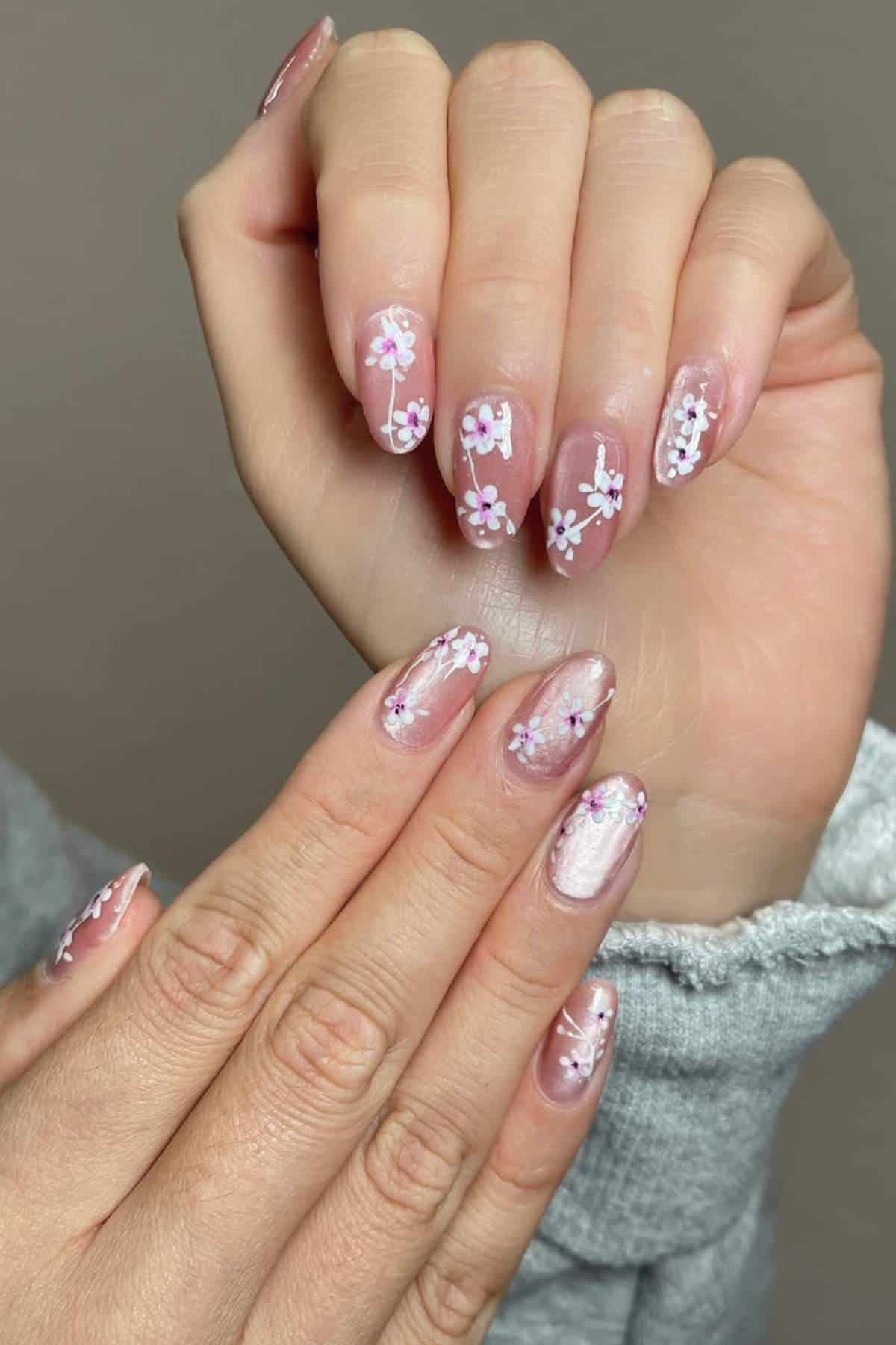 A person with medium-length, almond-shaped nails painted light pink with white and purple floral designs on each nail.