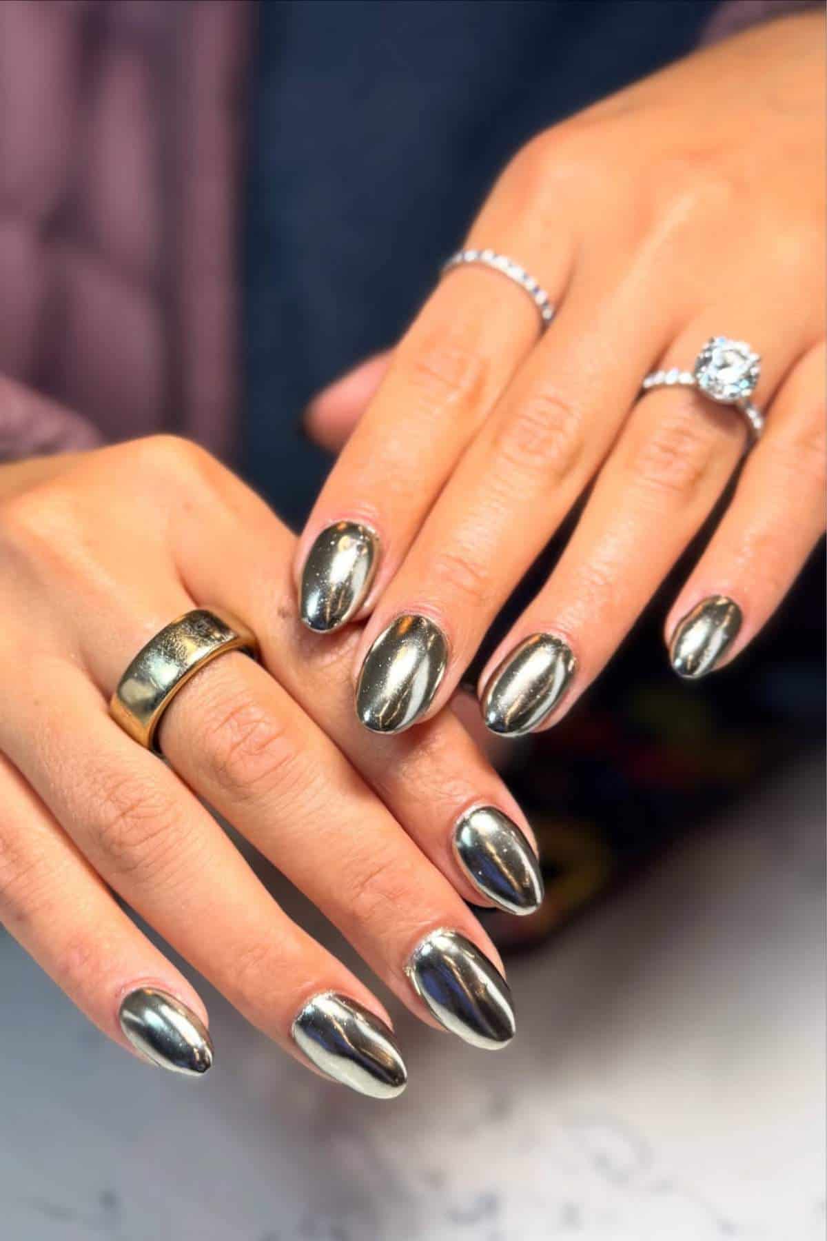 Close-up of hands with metallic silver almond-shaped nail polish, wearing a large gold band and a silver ring with a prominent round gemstone.