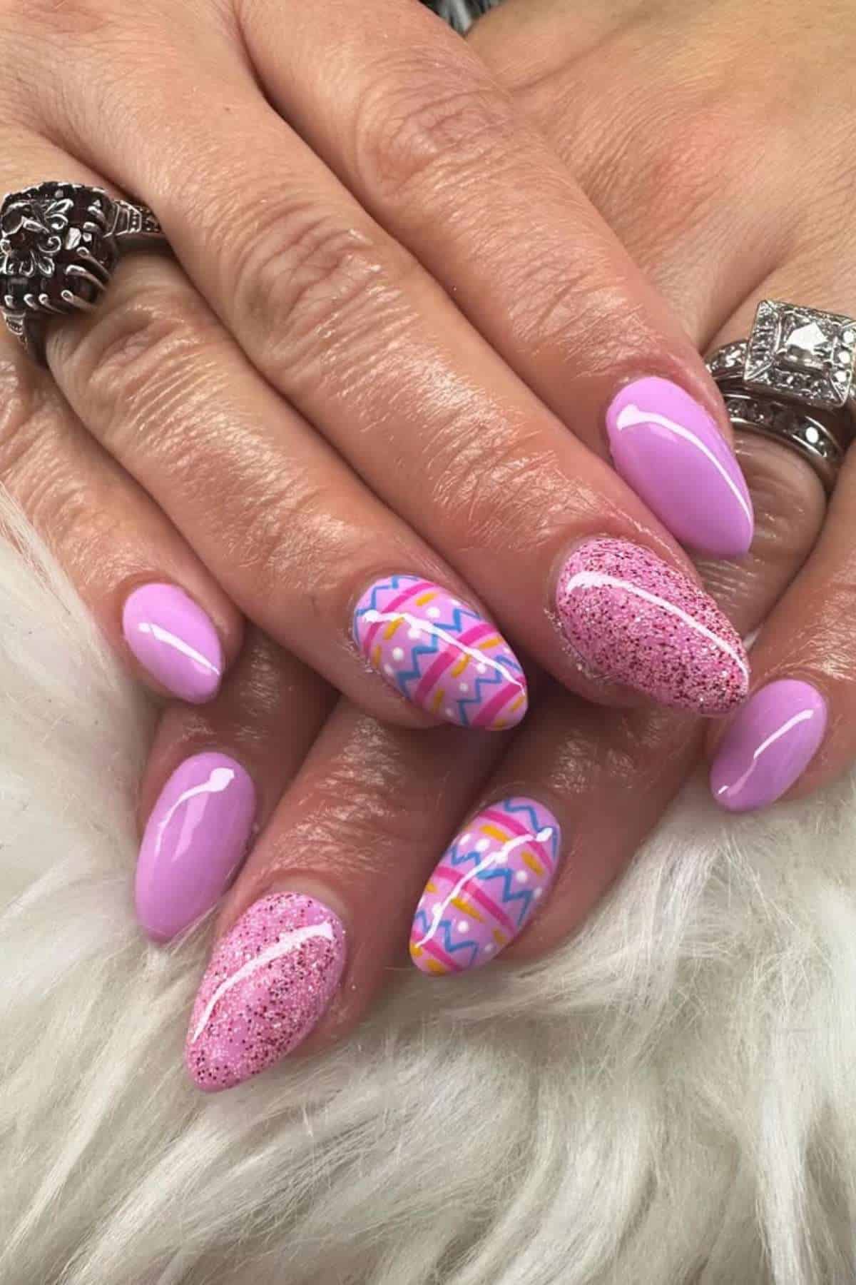 Hands with almond-shaped nails painted in pastel pink, featuring glitter accents and colorful geometric patterns on two nails, resting on a white fur surface.