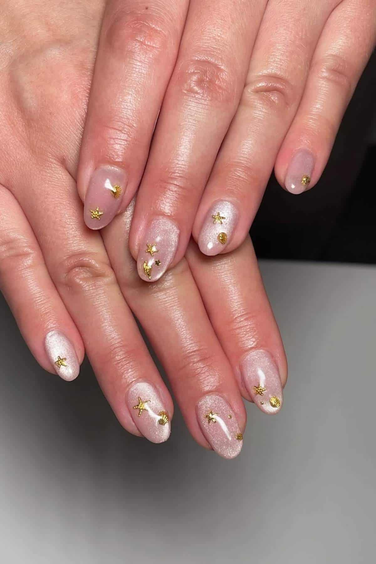 Hands with oval-shaped nails painted in a pale pink shade, featuring small gold foil accents on each nail.