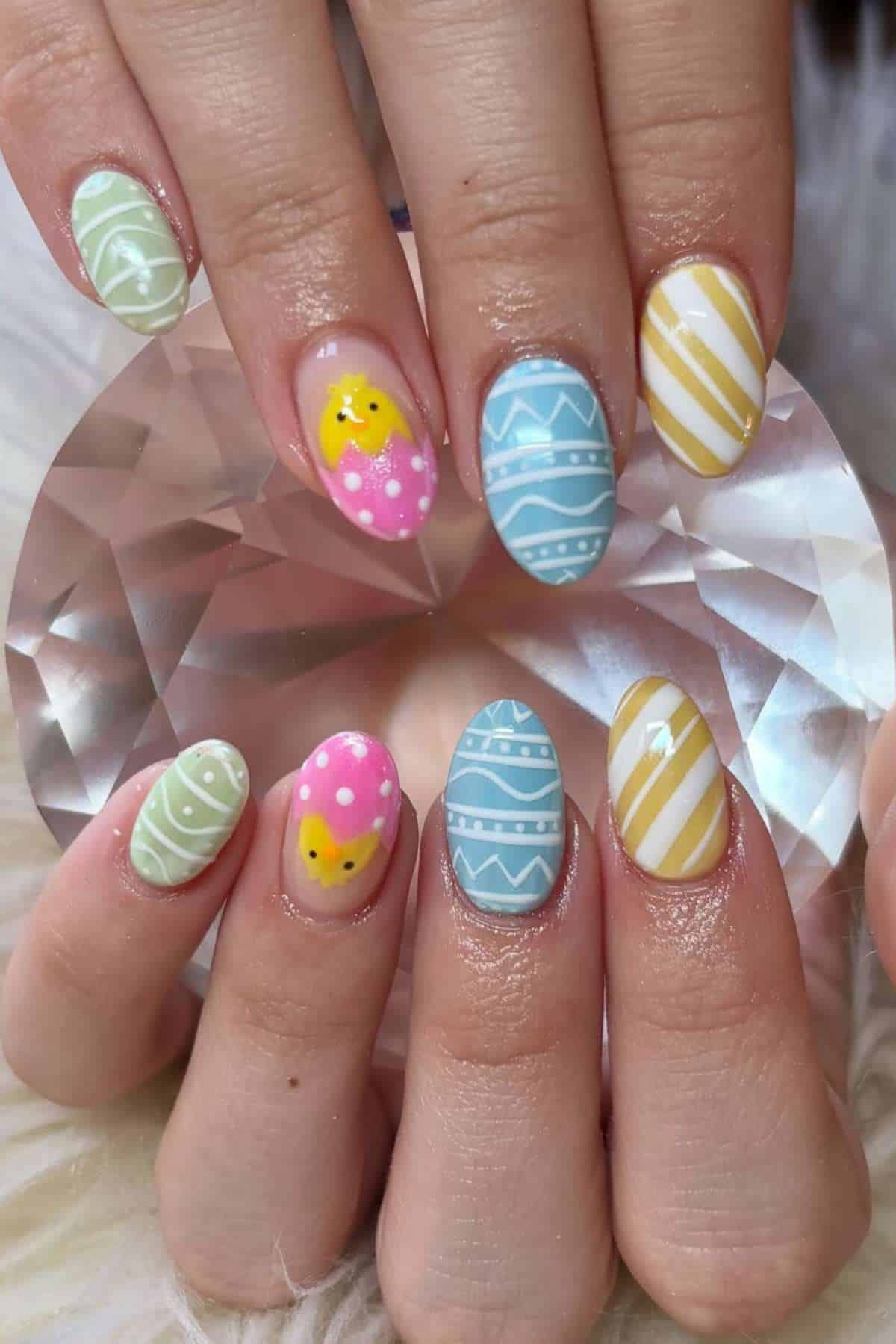 Hands with pastel-colored Easter-themed nail art, including egg patterns and a small yellow chick design, holding a clear faceted crystal.