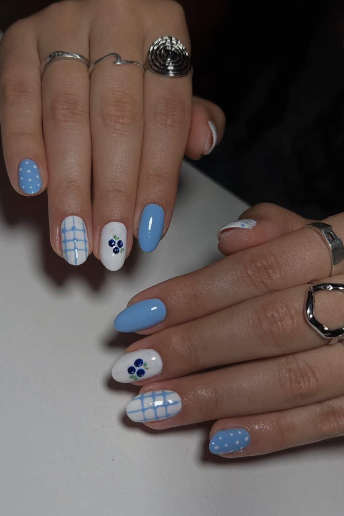 Hands with manicured nails featuring blue and white polish, polka dots, grid lines, and small flower designs; person wears silver rings on fingers.