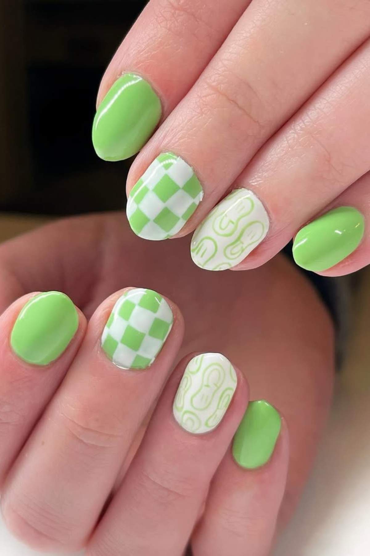 Close-up of hands with green and white nail art; some nails feature a checkered pattern and others have abstract line designs, while the rest are painted solid green.