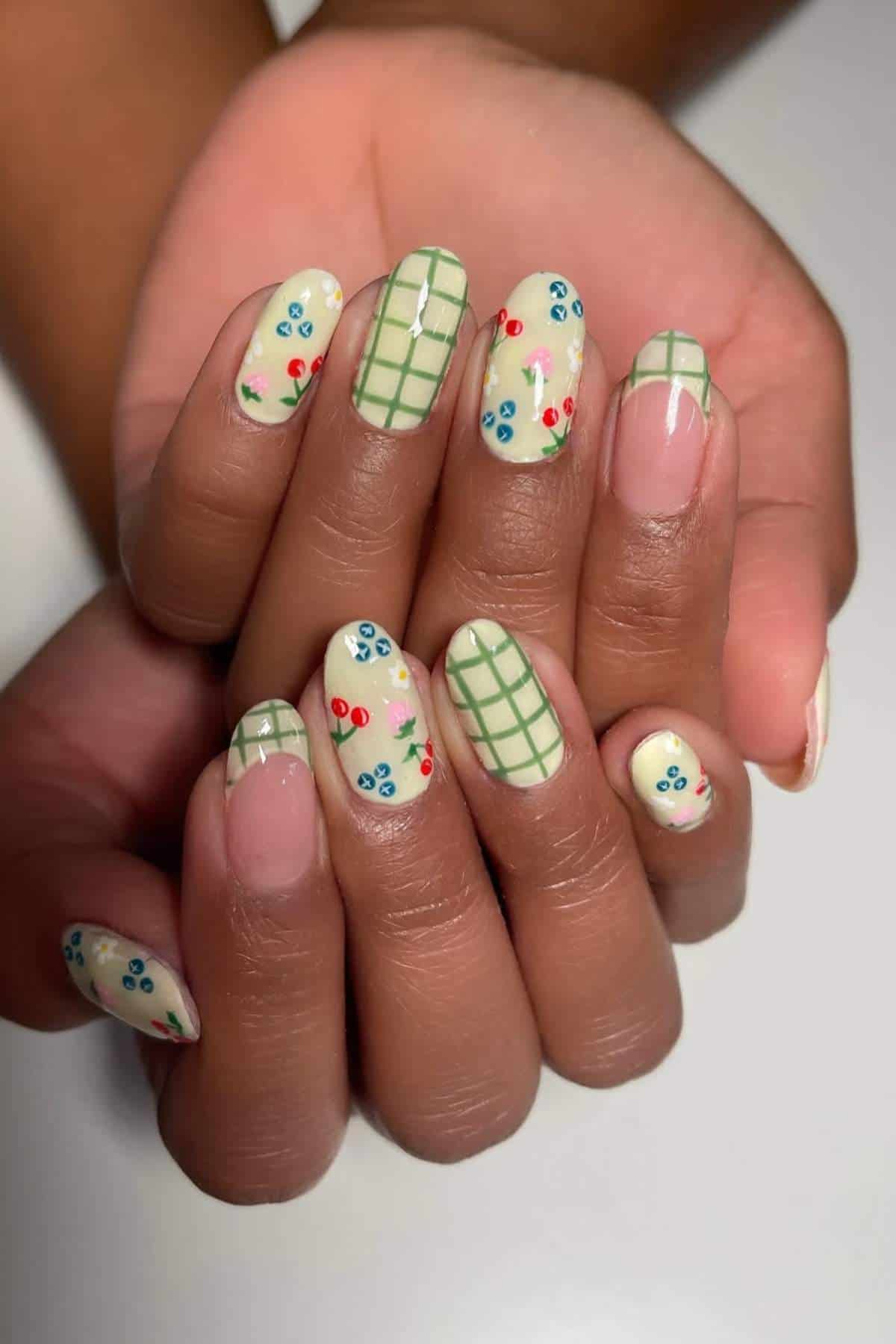 Hands with almond-shaped nails painted cream, featuring green grid lines and small red and blue floral designs as nail art.