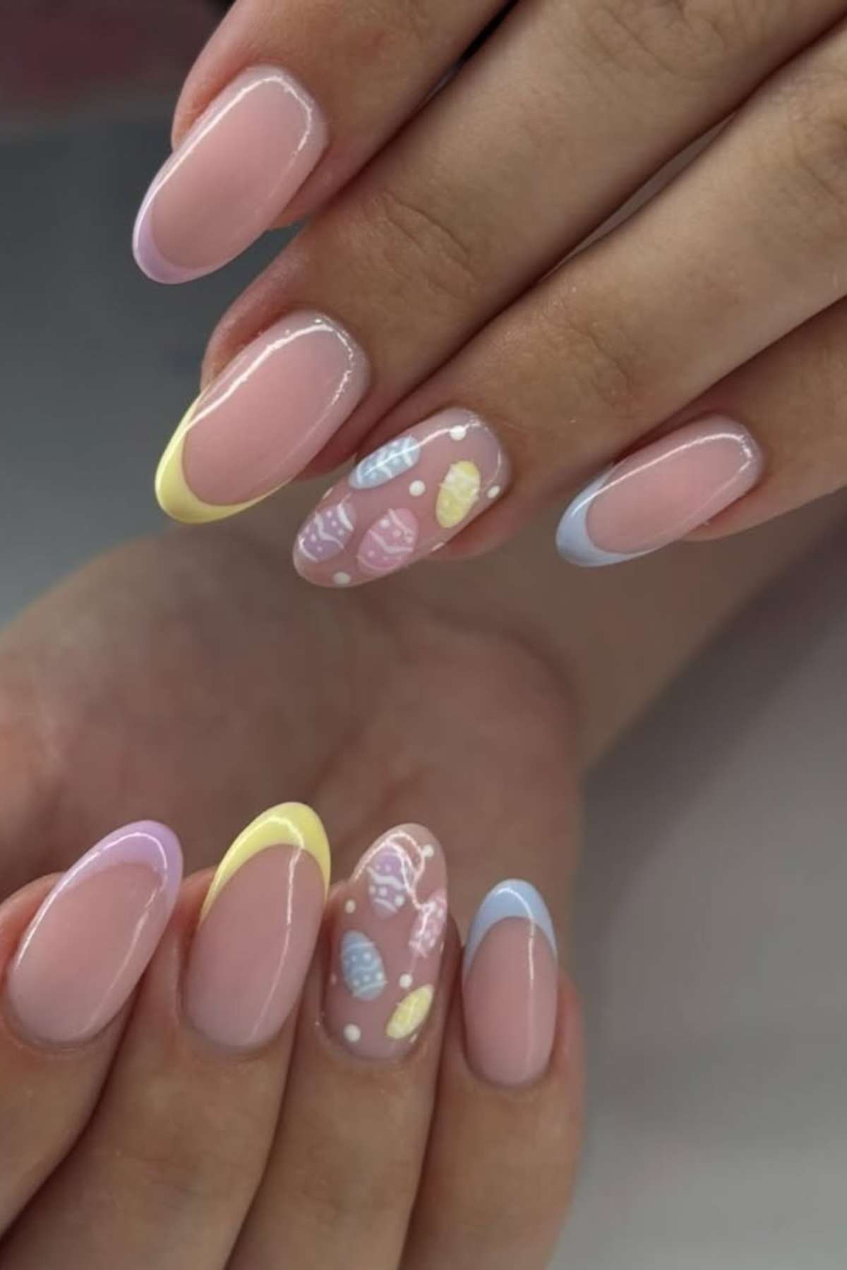 A hand with almond-shaped nails featuring pastel French tips and accent nails with white and pastel seashell designs.
