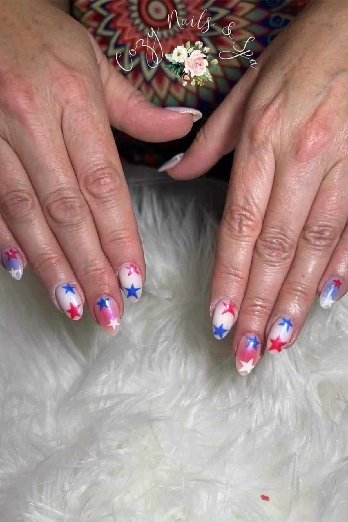 Hands with manicured nails featuring red, blue, and pink star designs on a white base, displayed on a white fuzzy surface.
