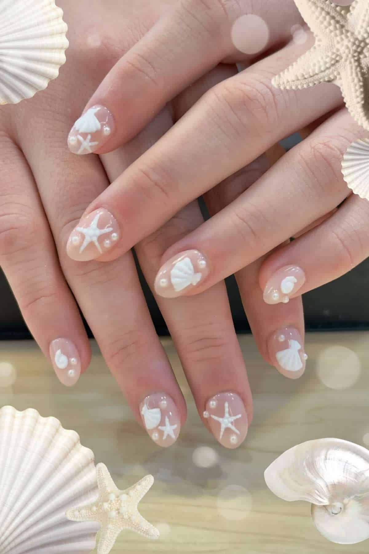 Close-up of hands showing fingernails with clear polish and white seashell, starfish, and pearl nail art designs, surrounded by illustrated seashell decorations.