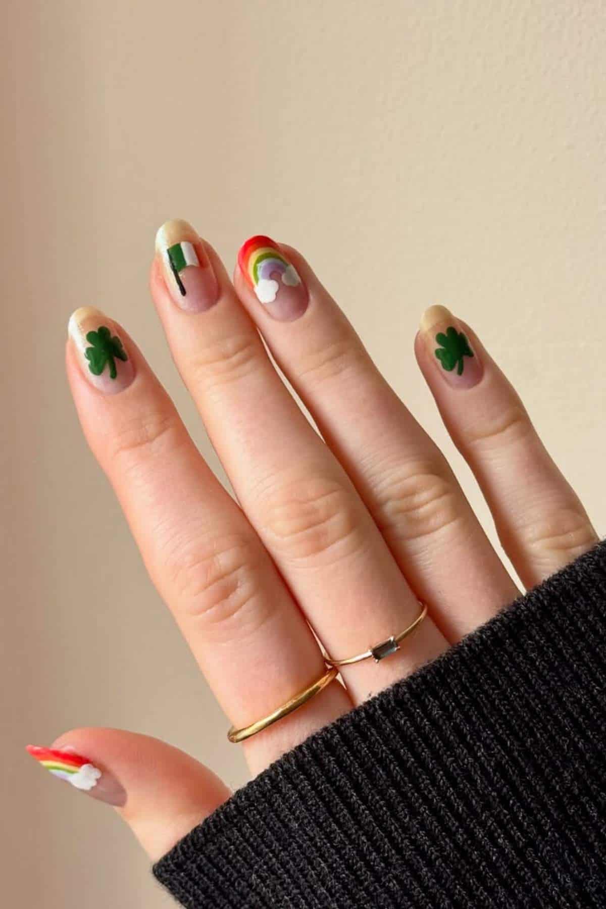 A hand with manicured nails featuring designs of shamrocks, the Irish flag, and rainbows, photographed against a plain light background.