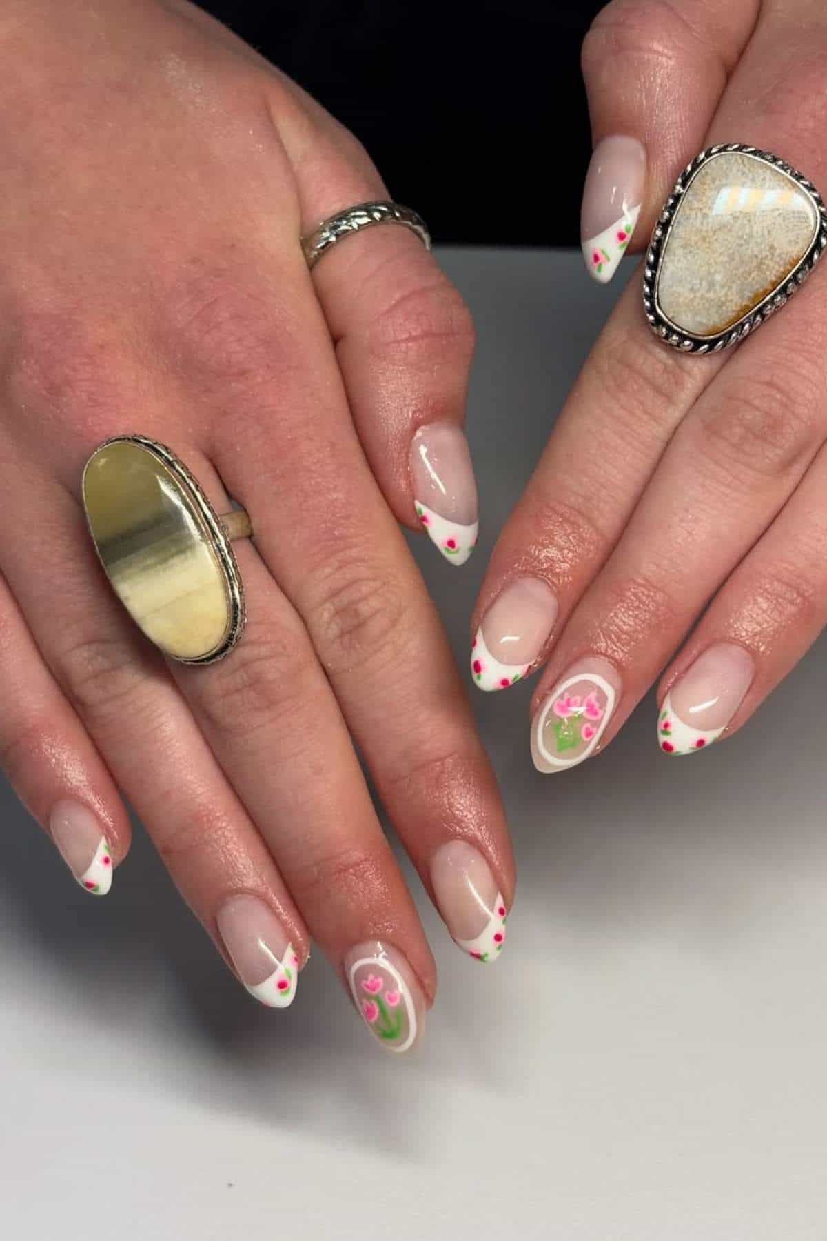 Hands with almond-shaped nails featuring a French manicure with white tips, small red dots, and pink floral designs; two fingers have large rings.