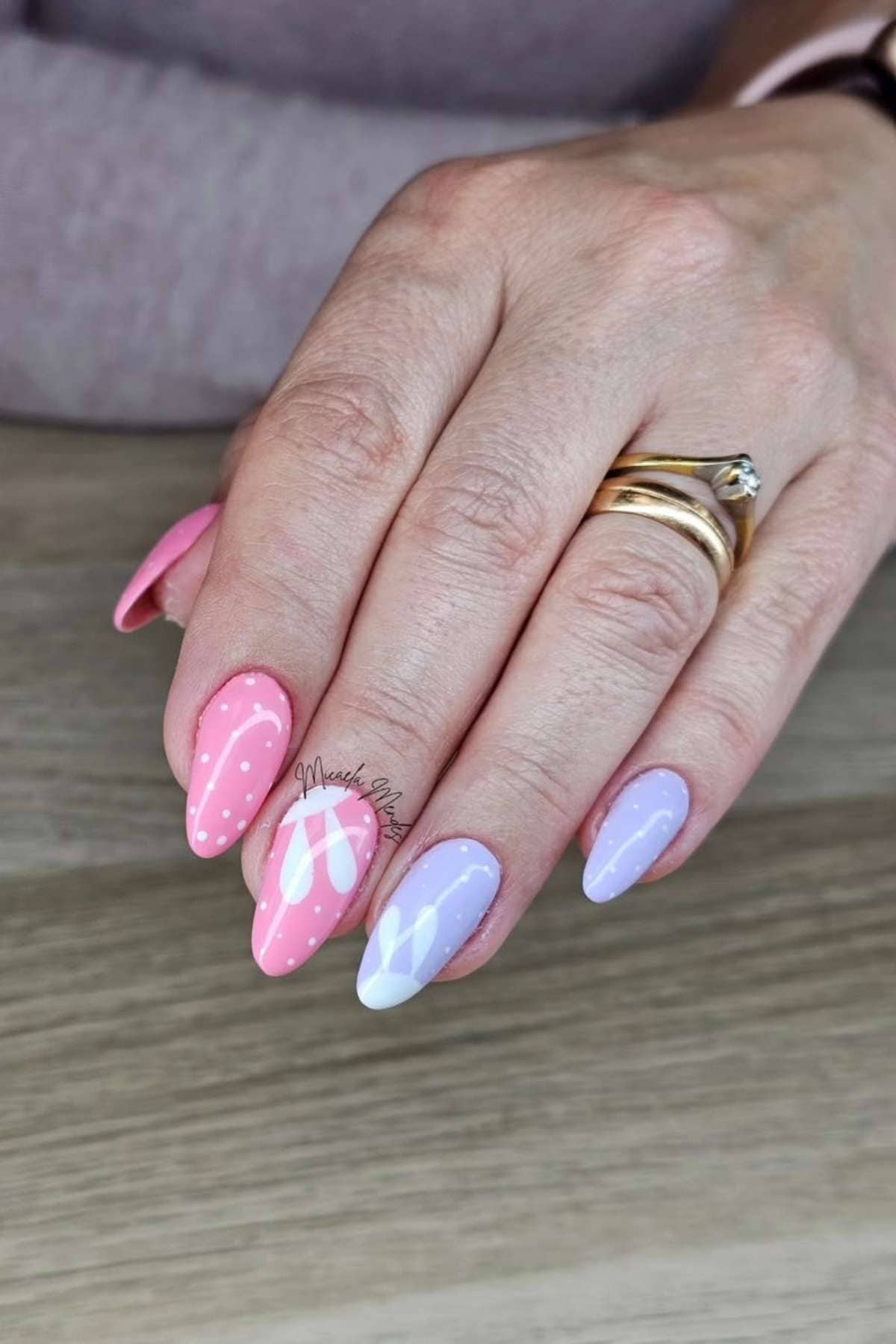 A hand with long, almond-shaped nails painted in pink and lavender with white polka dots and floral designs, resting on a wooden surface.