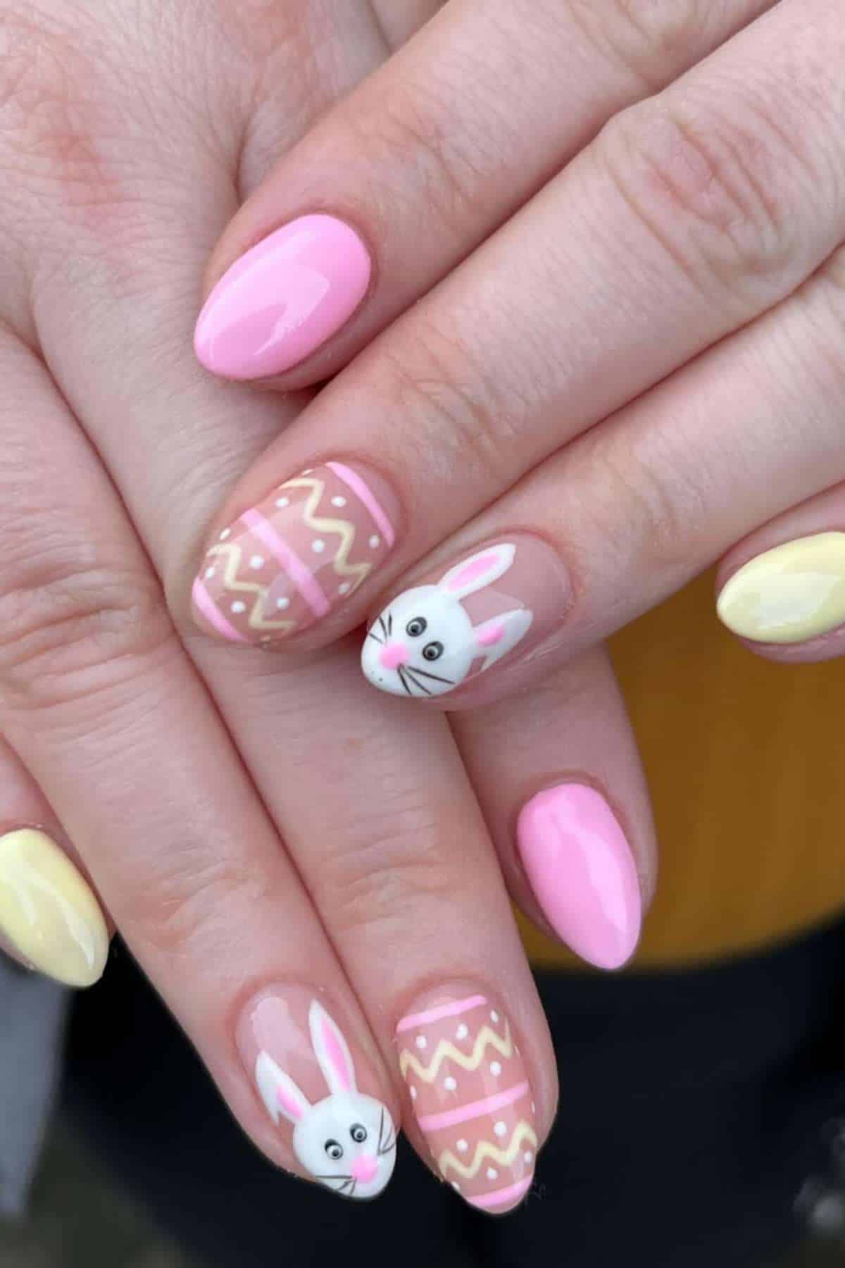 Hands with pastel pink and yellow manicure, featuring accent nails with Easter bunny faces and zigzag patterns.